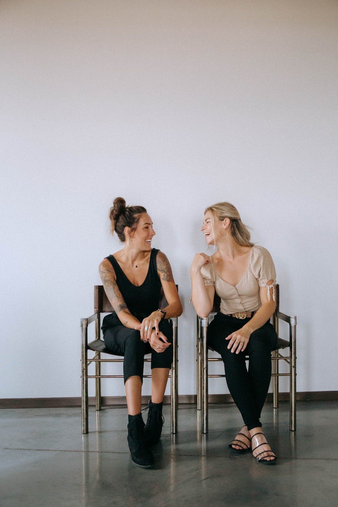 Two women sitting, smiling at each other. They're indoors near a white wall, on chairs. One has arm tattoos.