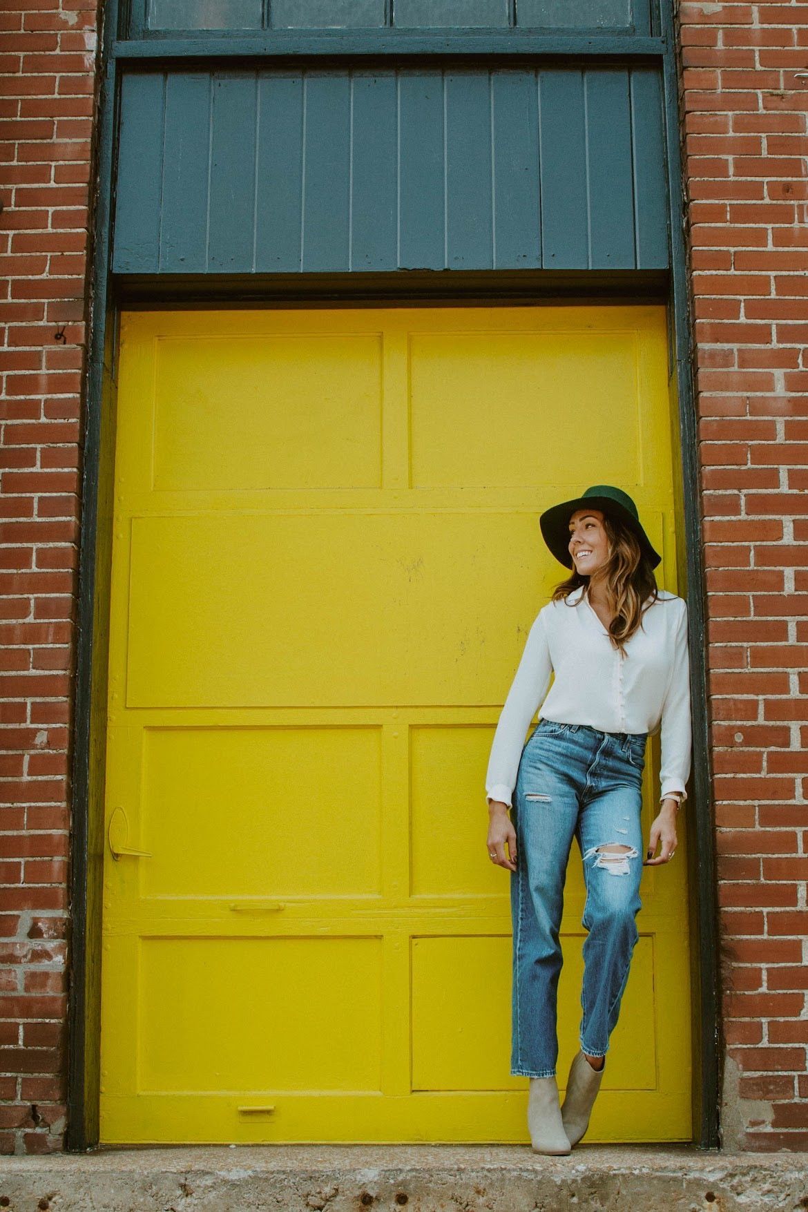 Woman wearing hat and jeans smiles near a bright yellow door against a brick wall.