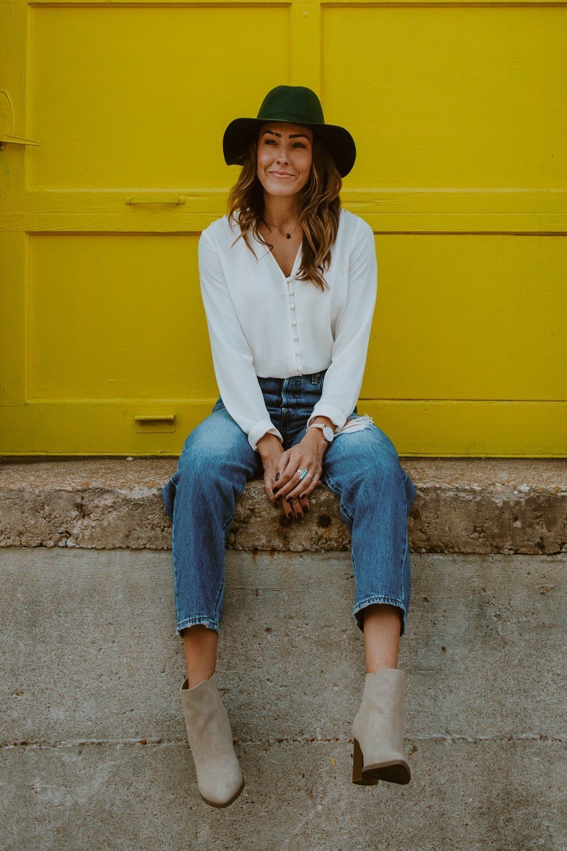 Woman in hat and jeans sitting in front of yellow door, smiling.