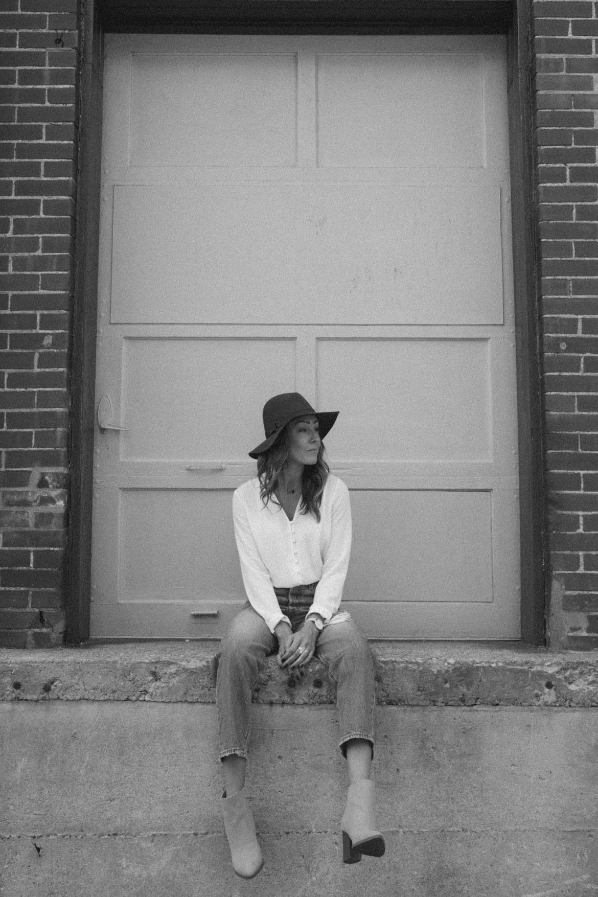 Woman in a hat and jeans sits on a ledge in front of a closed garage door. Brick building background.