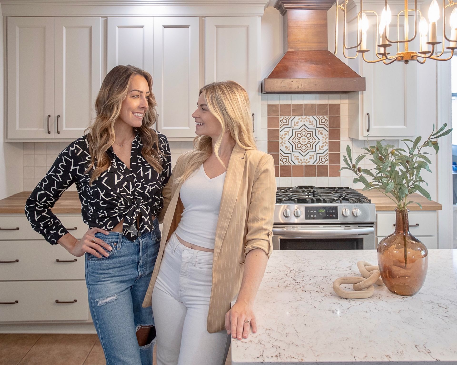 Two women in a kitchen, looking at each other. One wears a blazer, the other a patterned shirt. White cabinets and a gas range are in the background.