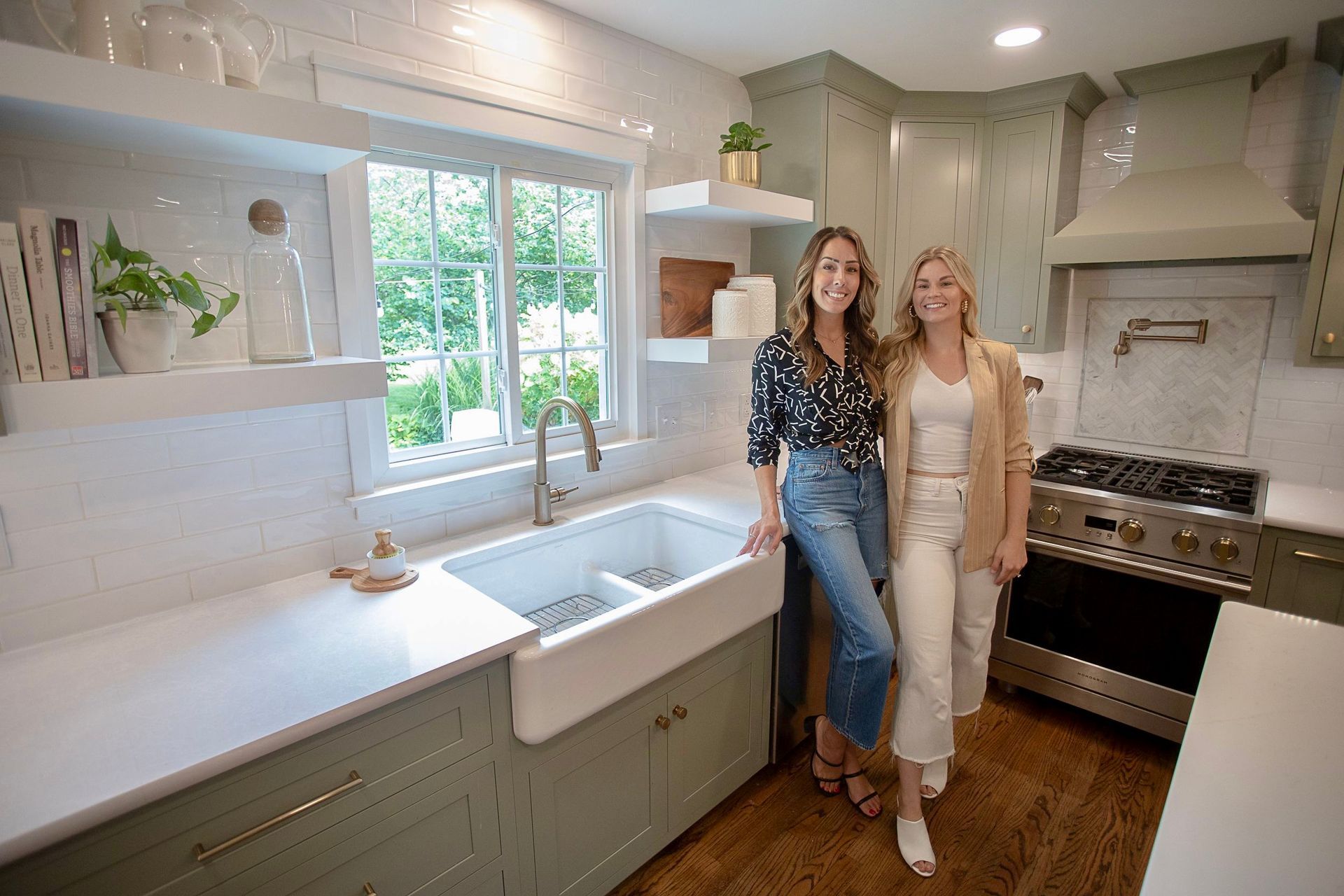 Two women pose in a green and white kitchen with a farmhouse sink and stainless steel appliances.