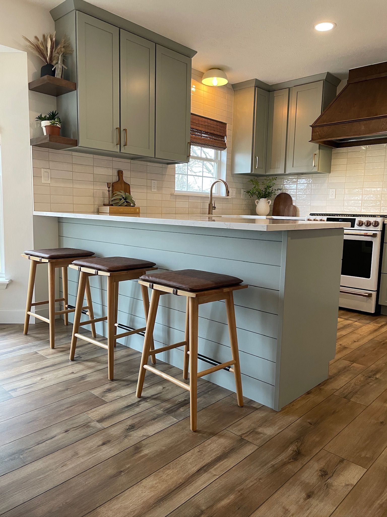 Kitchen with sage green cabinets, wood stools, and white tiled backsplash.