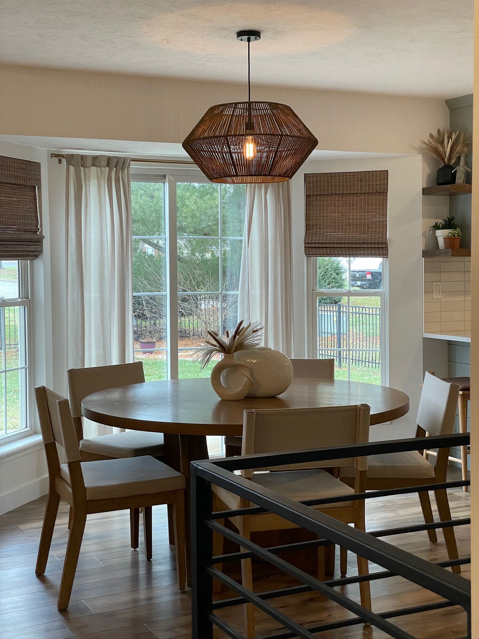 Dining room with round table, chairs, woven pendant light, and window with beige curtains and shades.