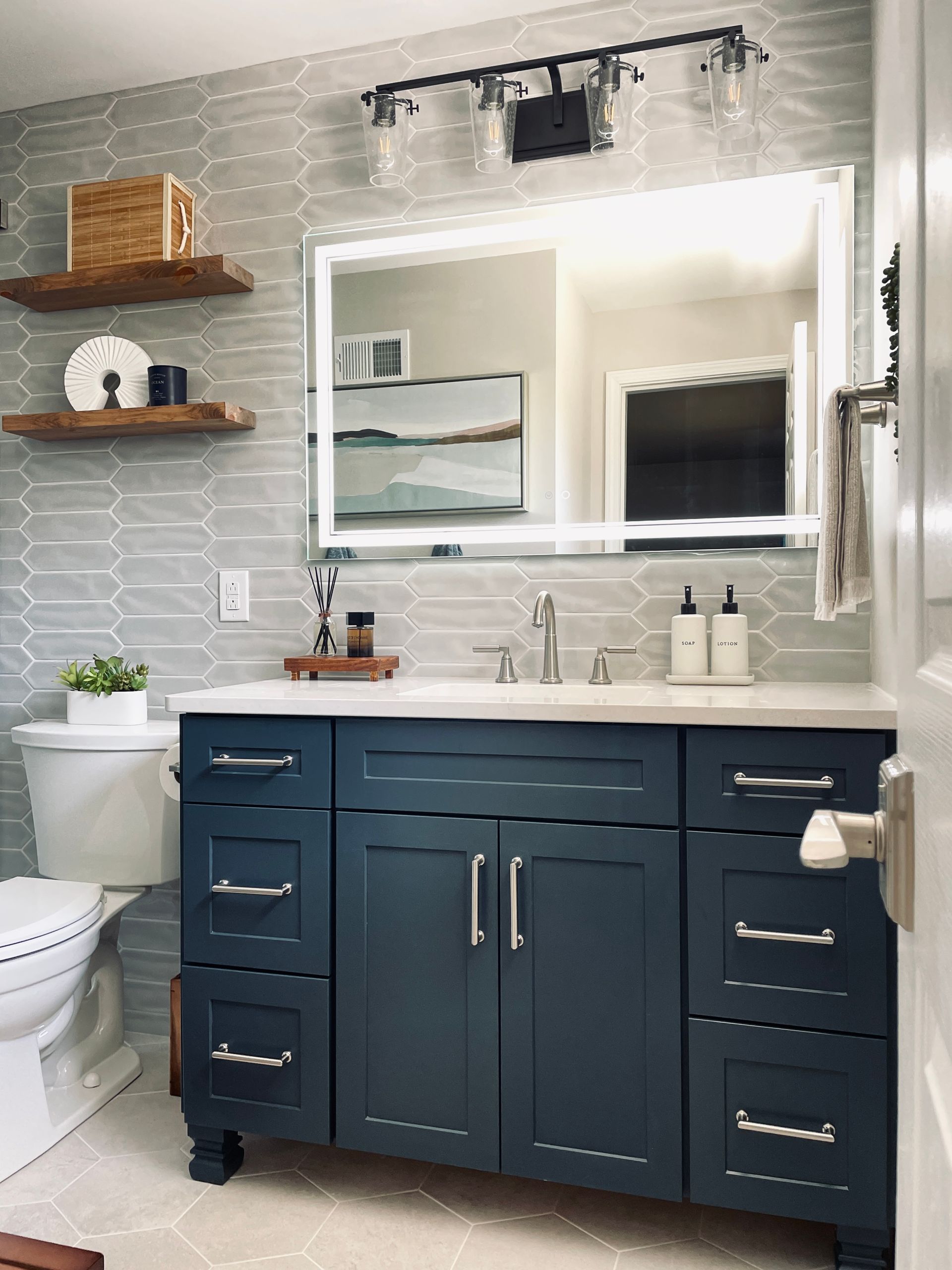 Blue bathroom vanity with white countertop and mirror with a backlit frame. Wooden shelves.