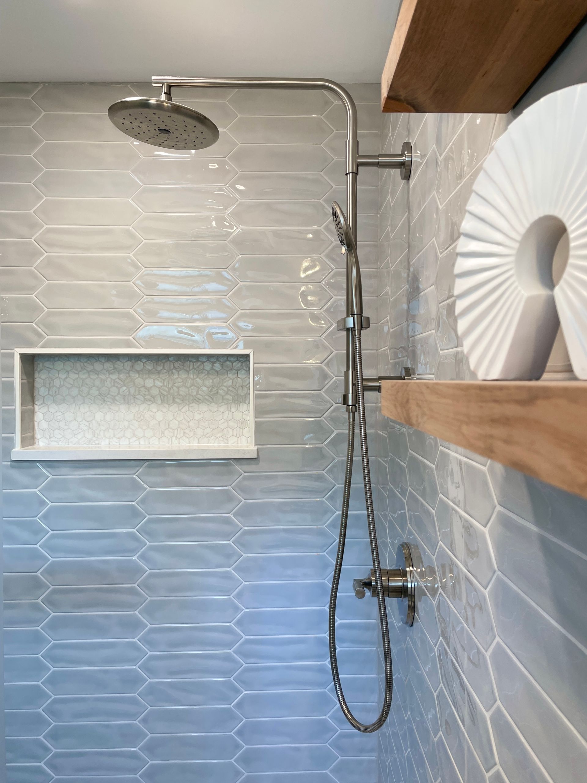 Shower with light blue, elongated hexagon tiles and chrome fixtures; wooden shelf with white art.