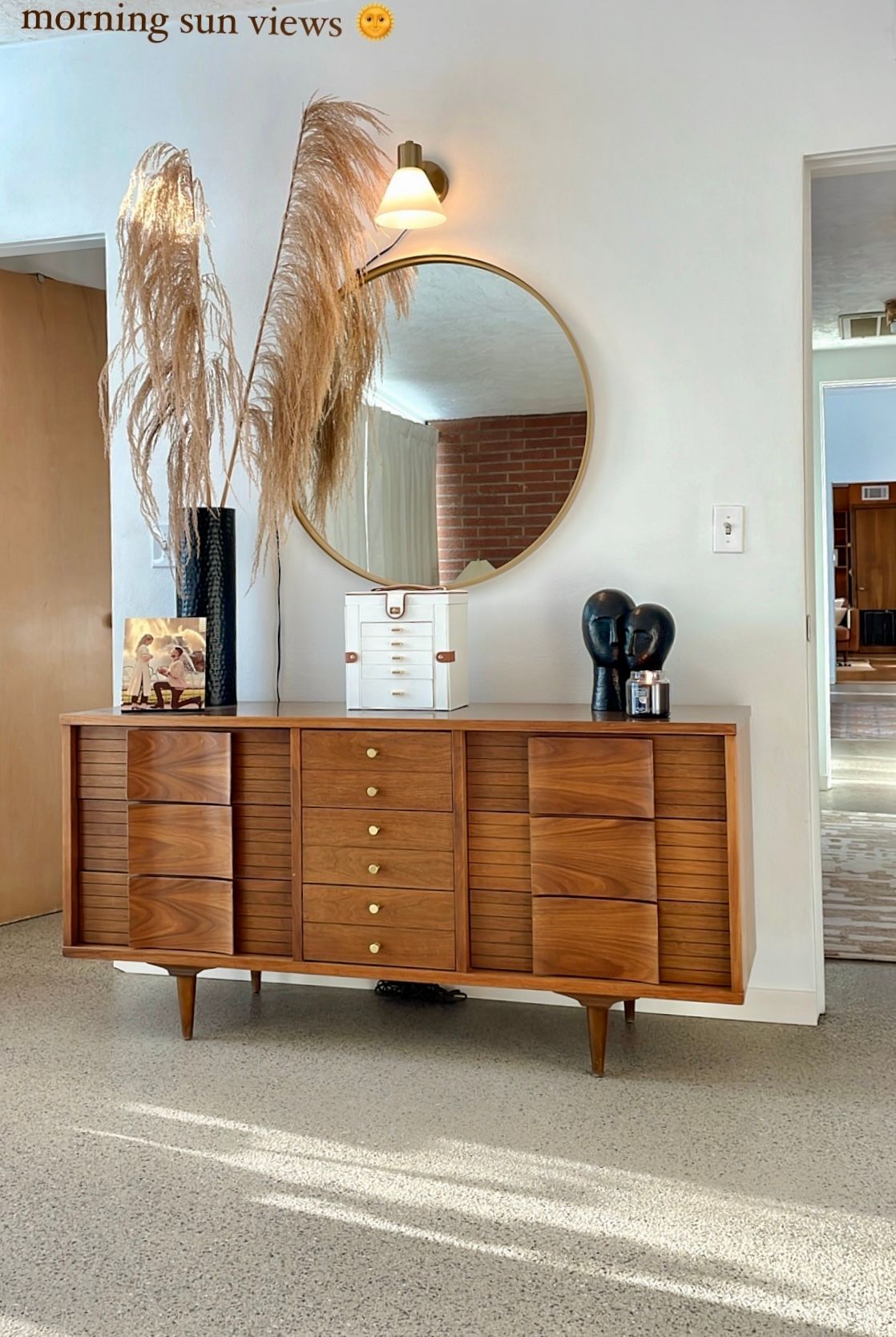 Mid-century modern style wood console with decor, round mirror on white wall, reed plumes, terrazzo floor.