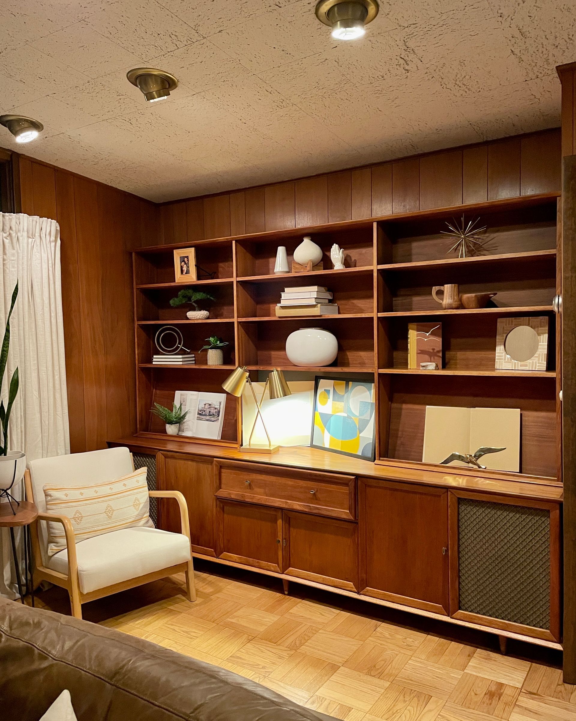 Wooden built-in shelving unit with decor, a beige armchair, and a glimpse of a couch in a cozy room.