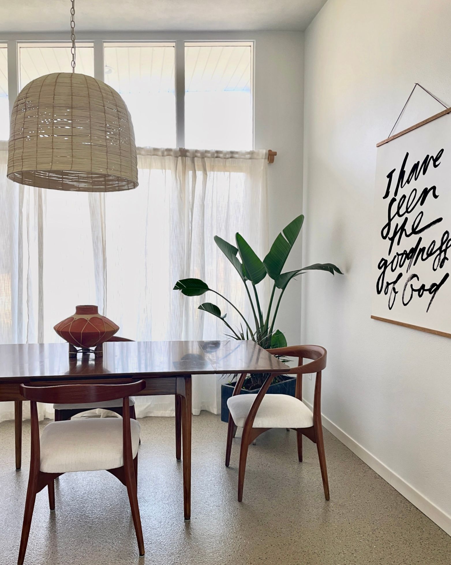 Dining room with a wooden table, chairs, and hanging pendant light. A plant and artwork decorate the room.