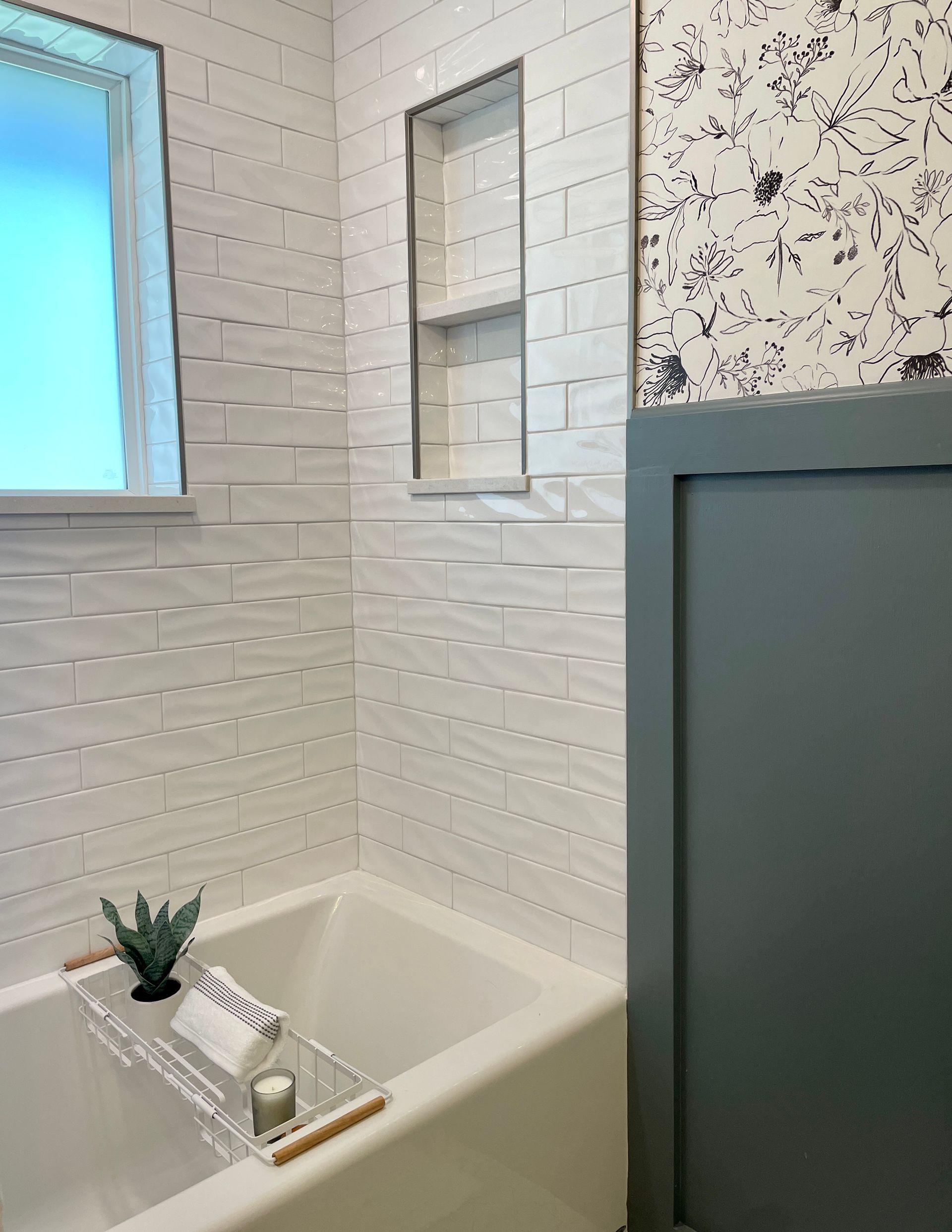 White-tiled bathroom with a bathtub and built-in shelf. Decorative wall paneling and floral wallpaper.