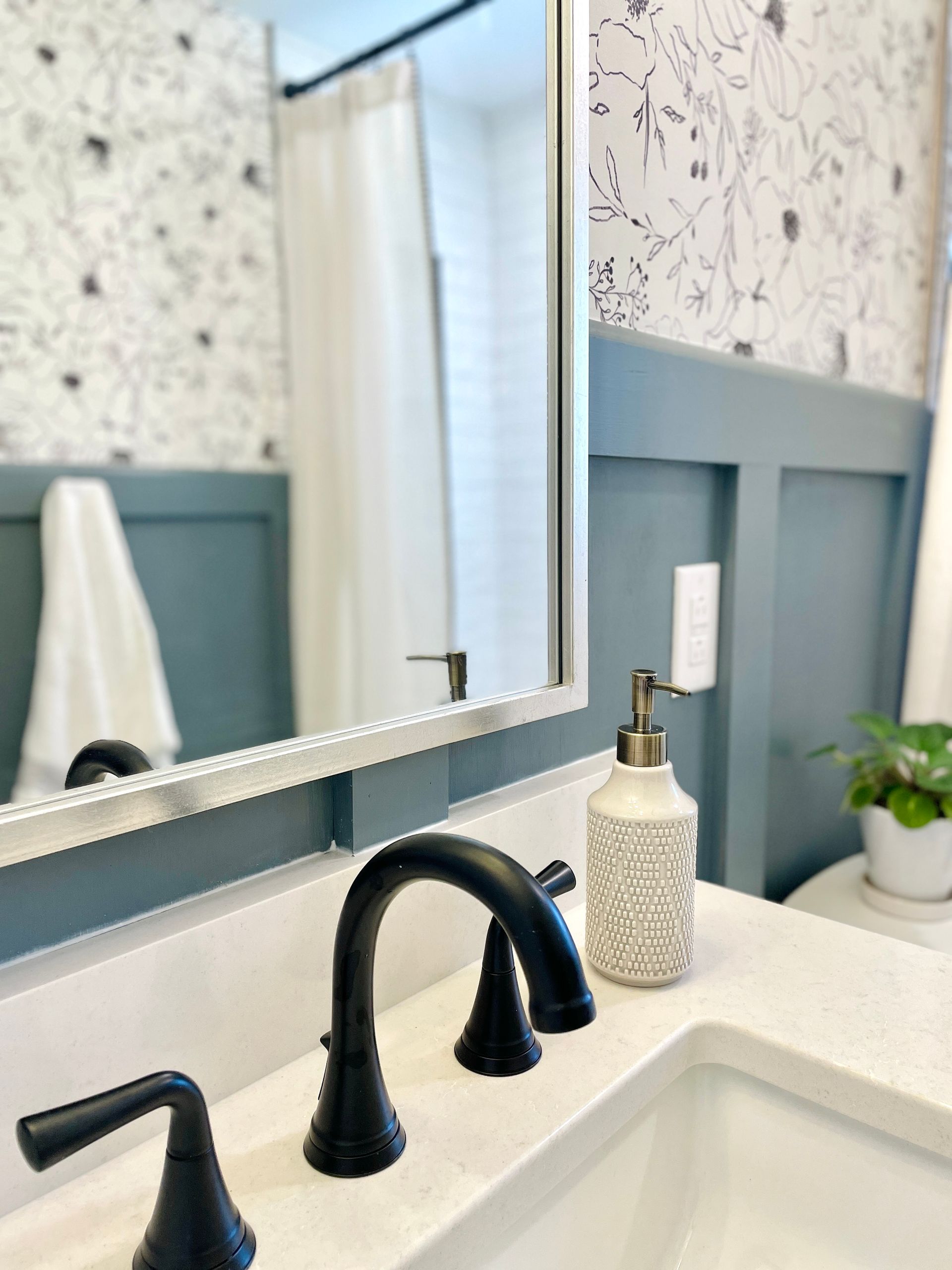Bathroom sink with black faucet, white countertop, blue paneled wall, and floral wallpaper.