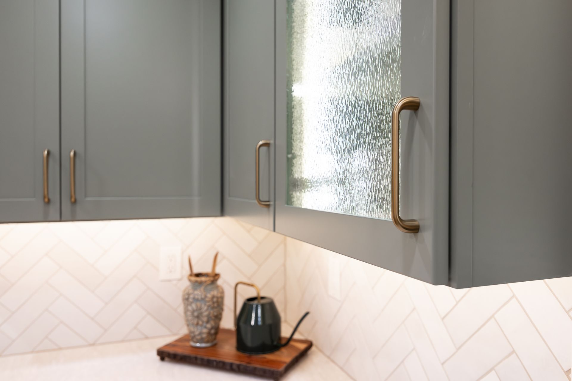 Gray kitchen cabinets with gold hardware and a glass front, above a white tiled backsplash.