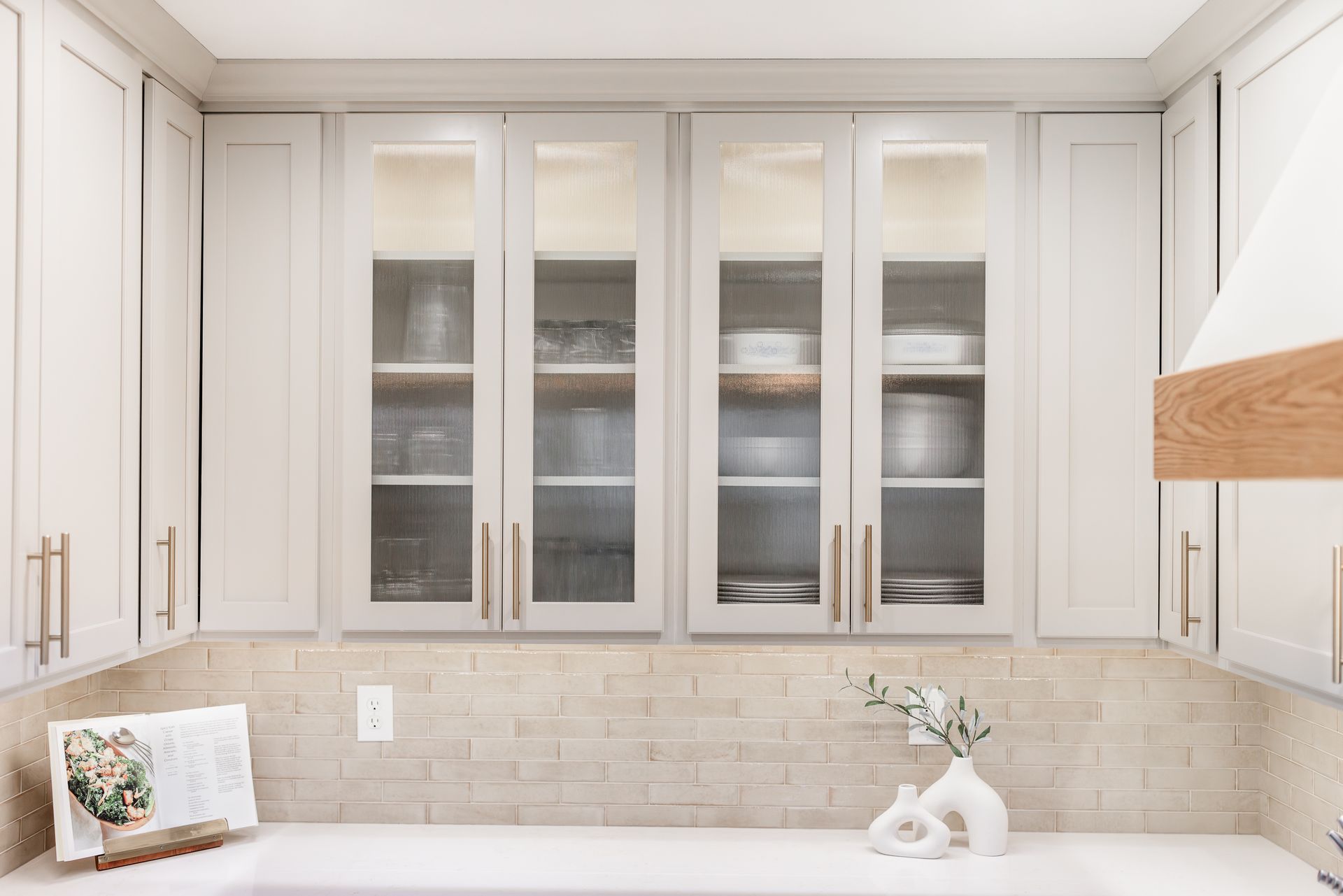 White kitchen cabinets with glass doors, displaying dishware. Beige brick backsplash, white countertop.