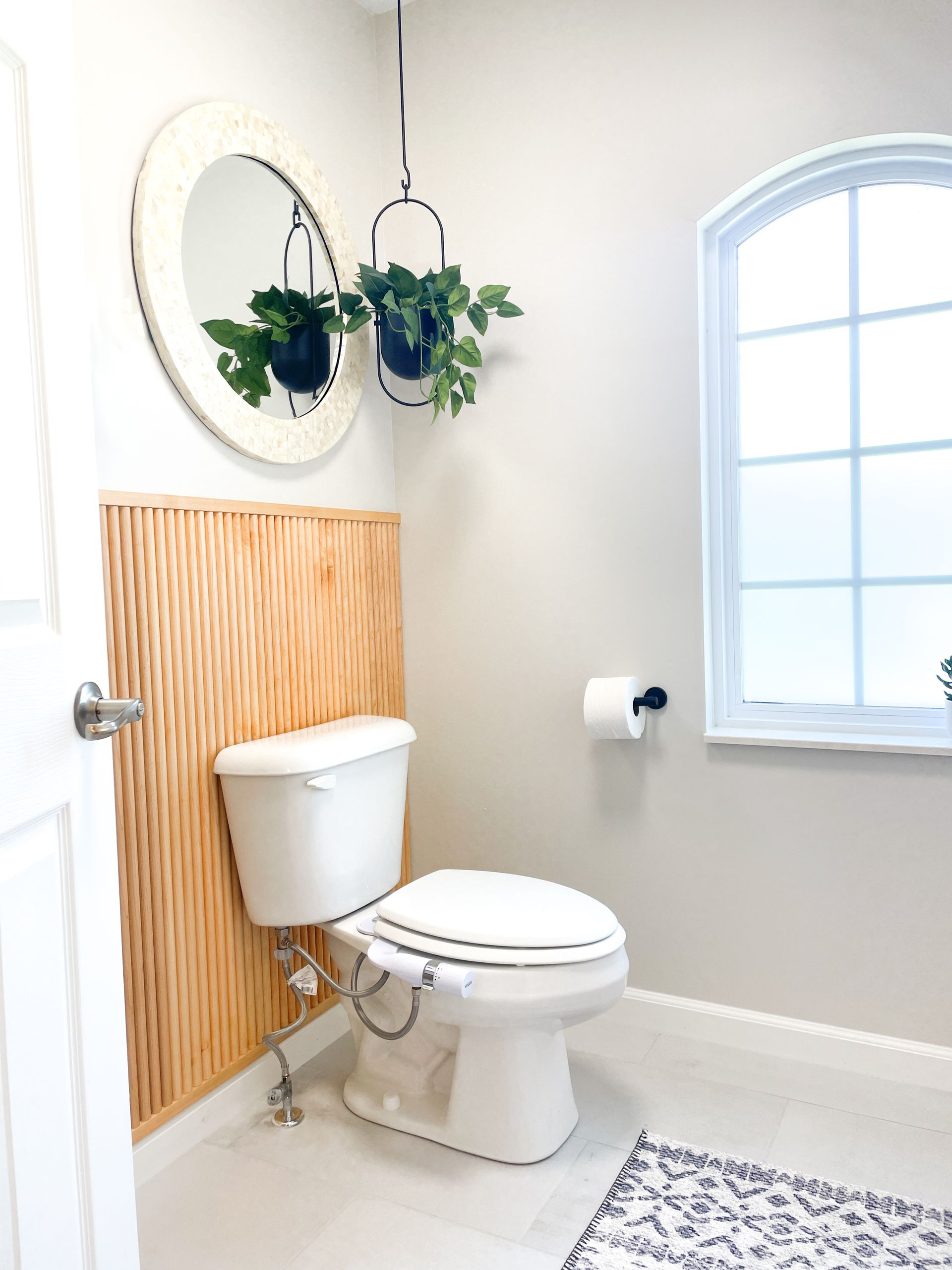 A bathroom with a toilet, mirror, plants, and a window. Light grey walls, wooden accent.