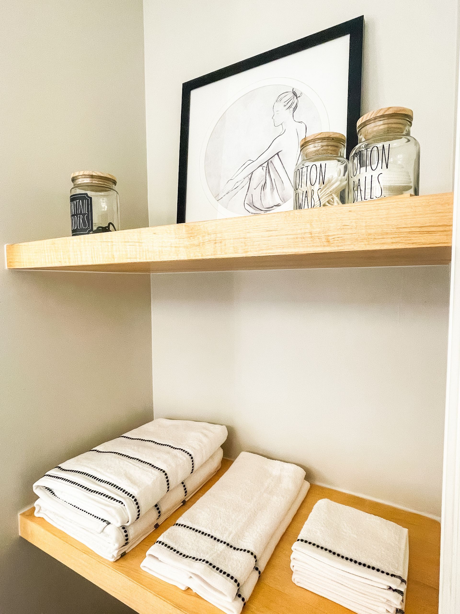 Two wooden shelves in a corner. The top shelf has jars and framed artwork. The bottom shelf holds folded white towels.