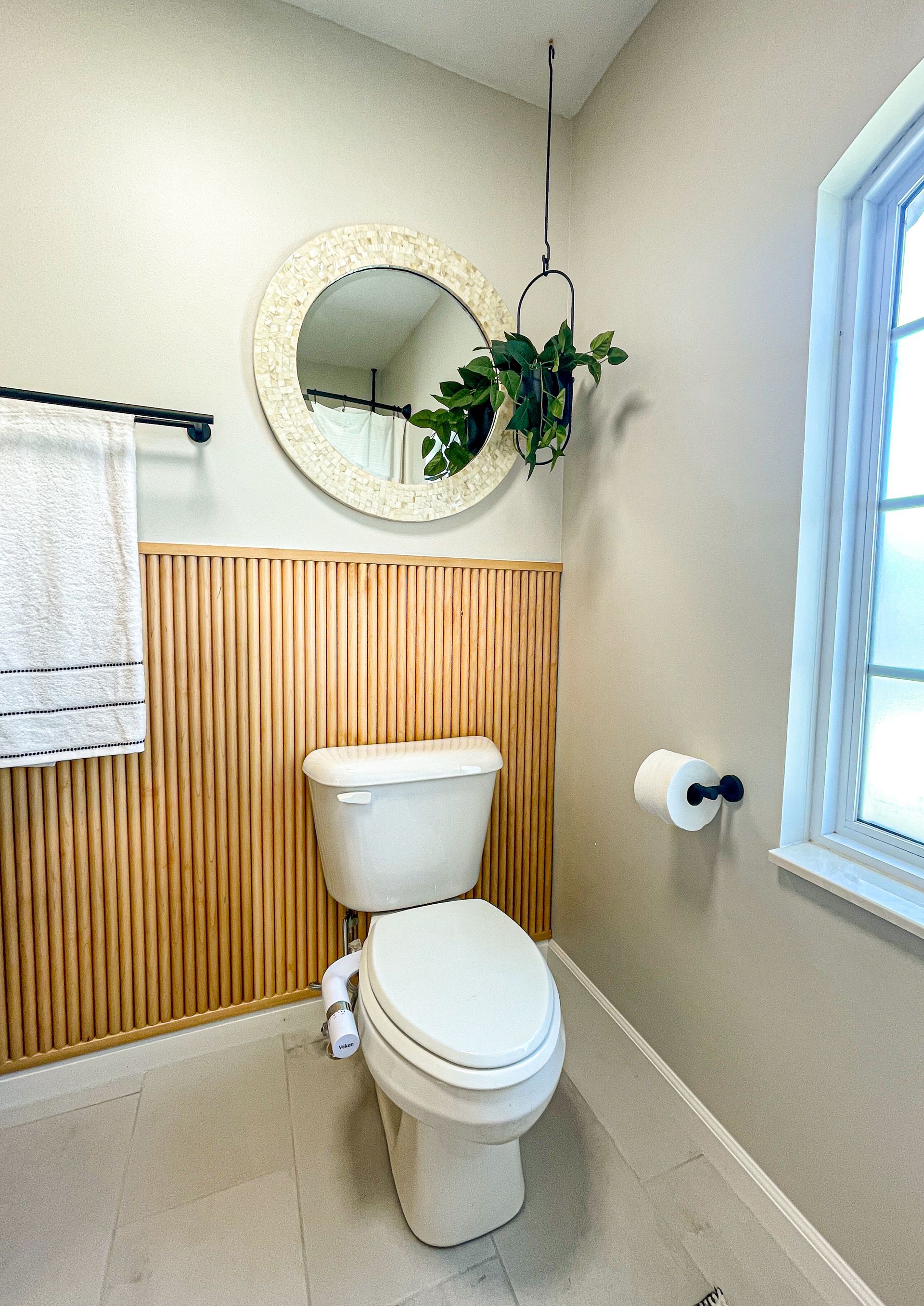 Bathroom with a wood panel wall, toilet, round mirror, and a hanging plant.