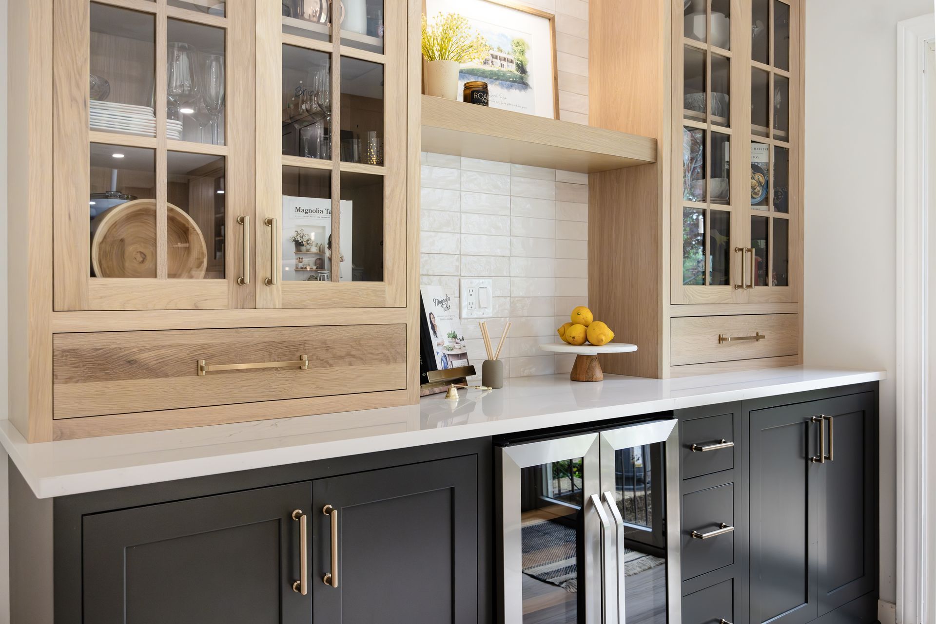 Built-in bar with light wood cabinets, white countertop, and a dark base. Includes a wine fridge and decor.