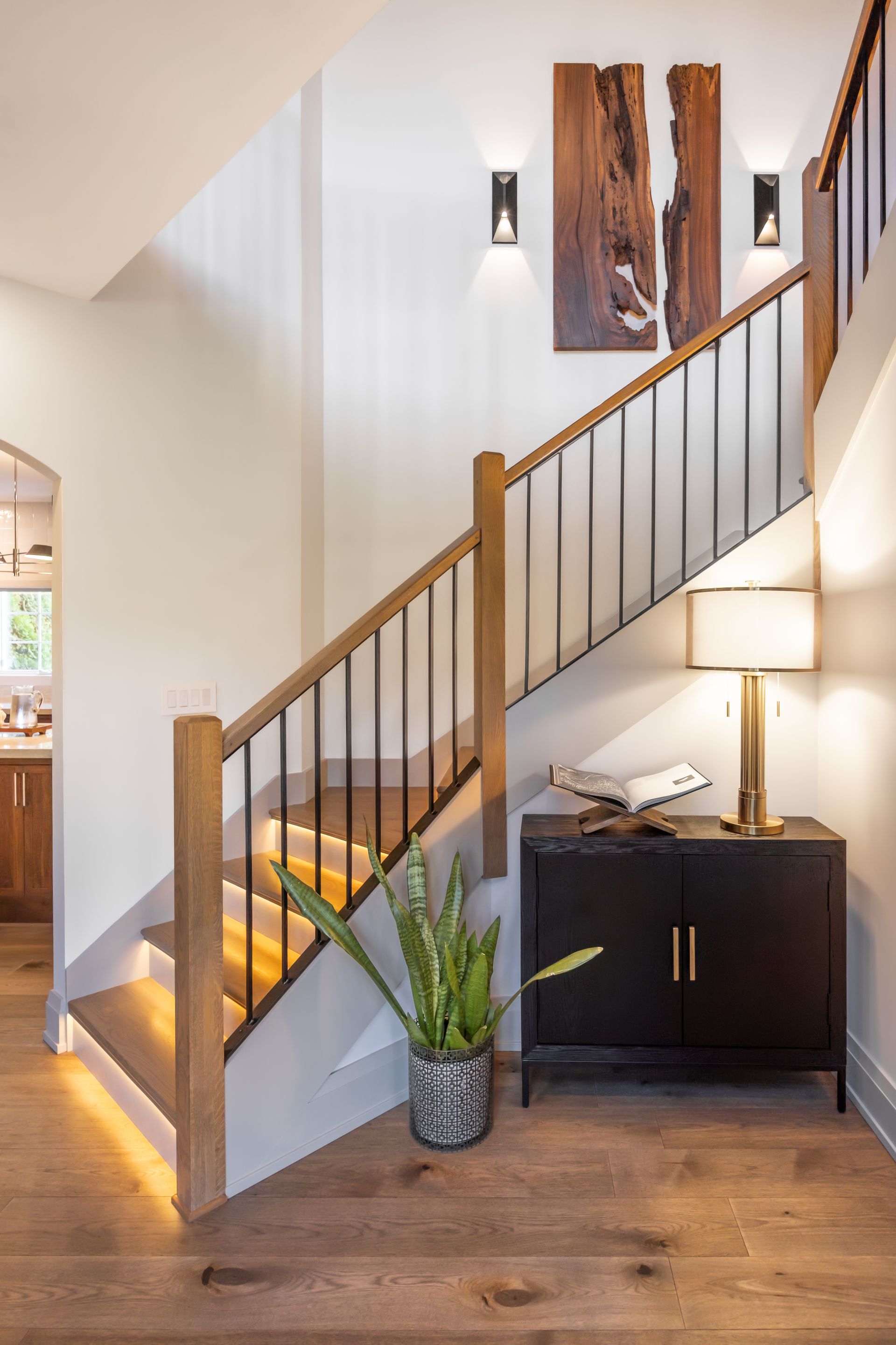 Custom staircase with LED lighting, light wood steps, black wrought iron railing, white or light gray walls, contrasting trim, leading to a lower level, custom wood wall art, modern wall sconces, modern transitional design, remodel in Edwardsville, IL
