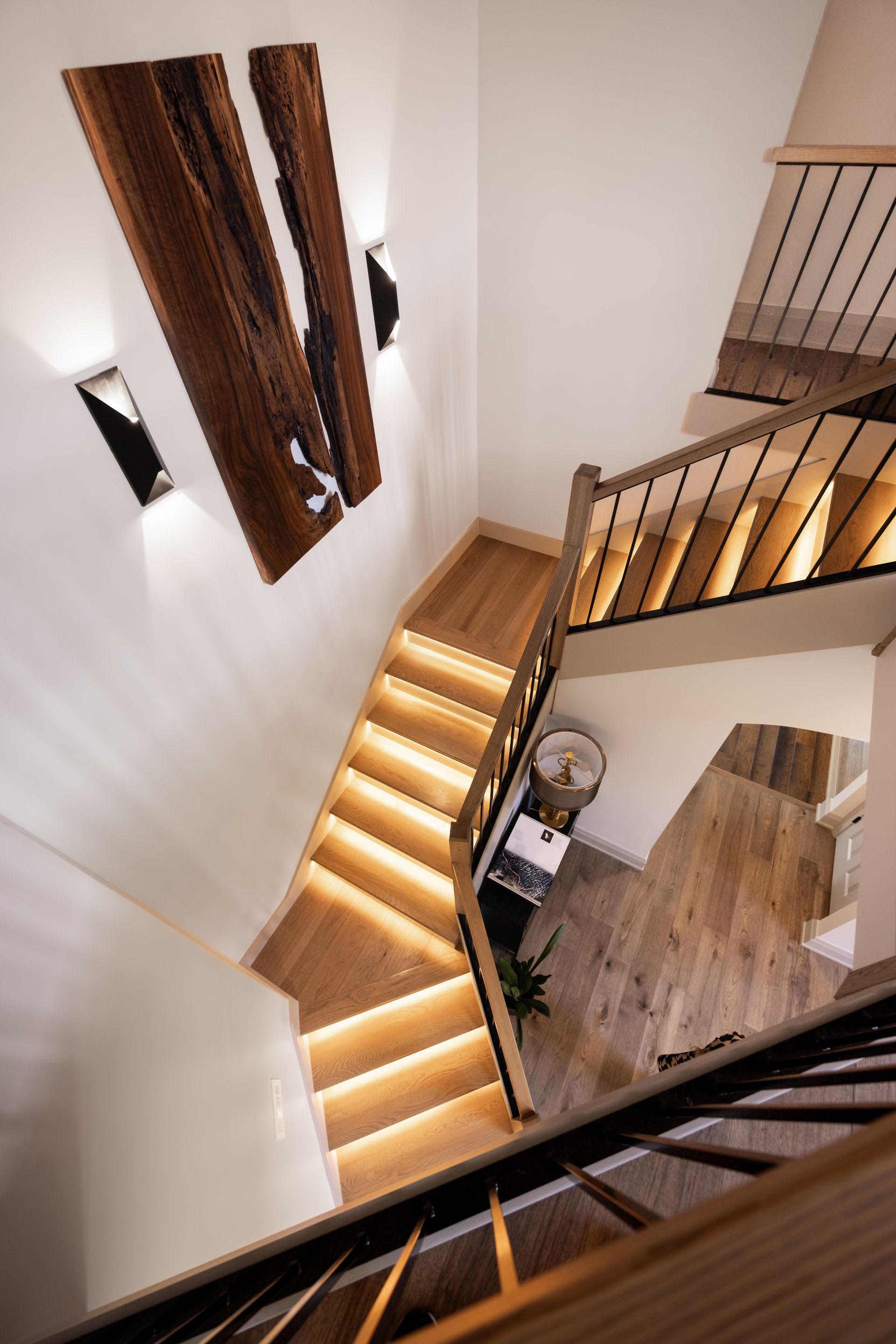 Custom staircase with LED lighting, light wood steps, black wrought iron railing, white or light gray walls, contrasting trim, leading to a lower level, custom wood wall art, modern wall sconces, modern transitional design, remodel in Edwardsville, IL