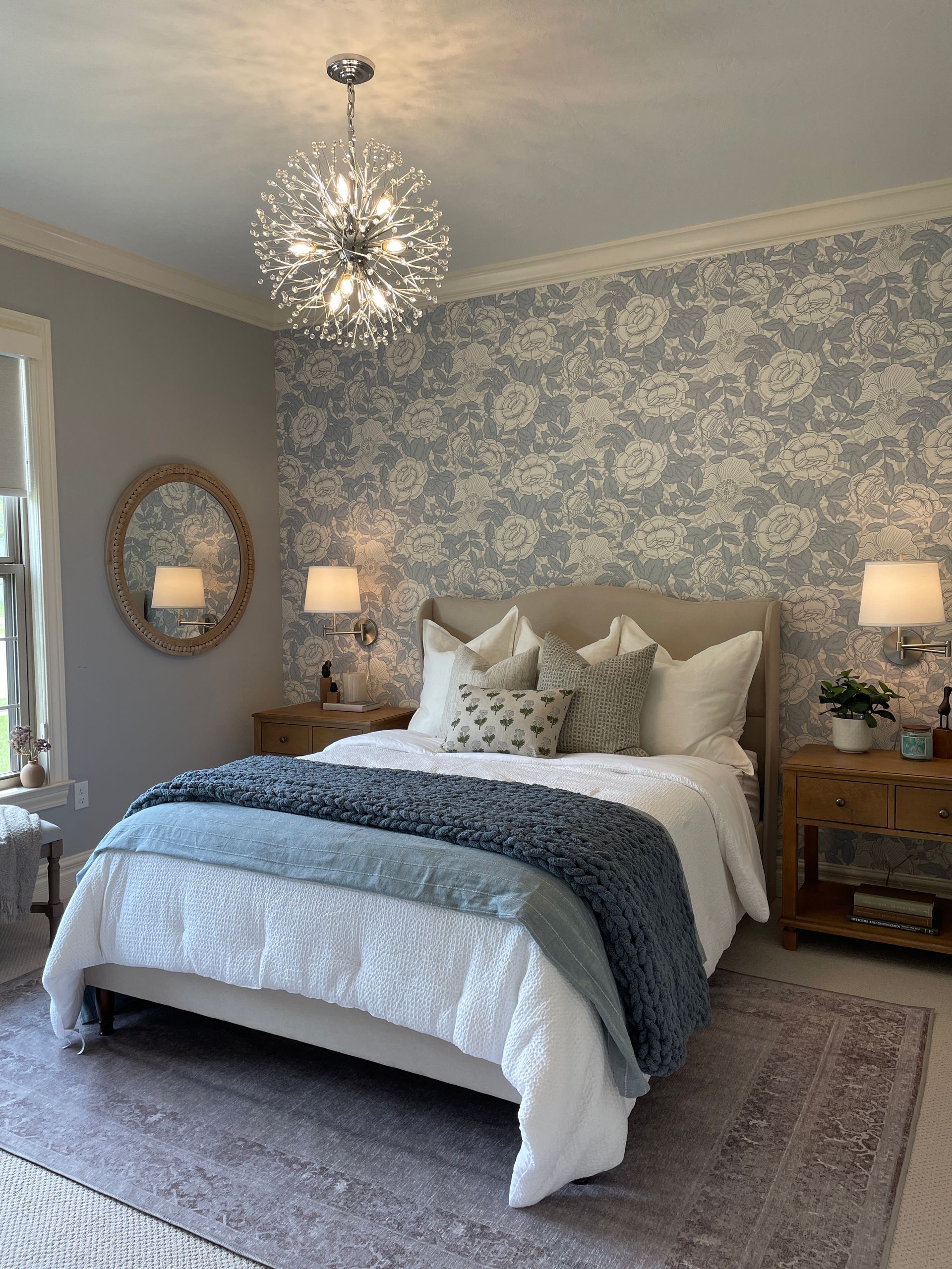 Bedroom with floral wallpaper, bed with blue accents, round mirror, and a sparkling chandelier.