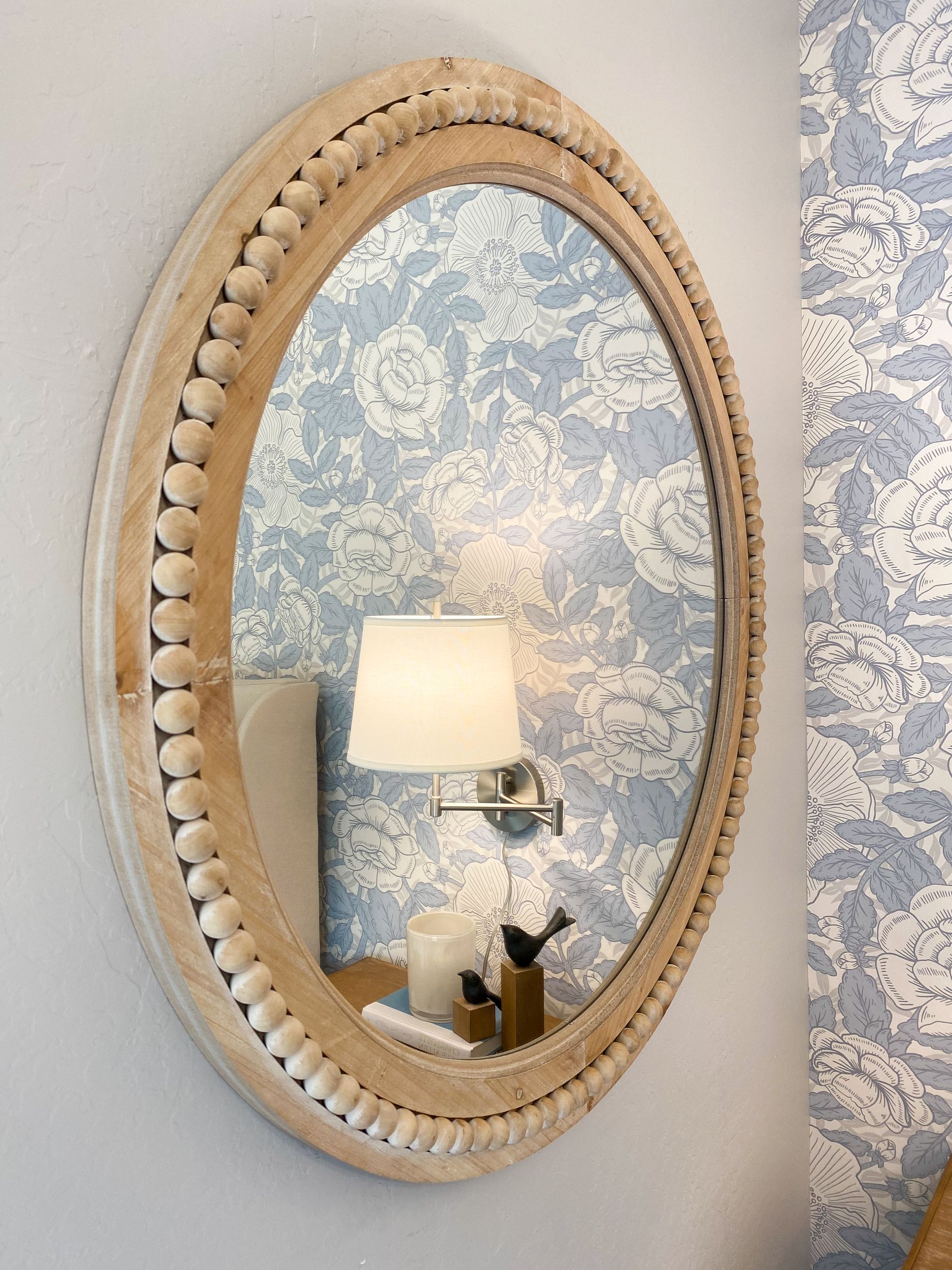 Oval mirror with beaded wood frame on a light blue floral wallpapered wall.