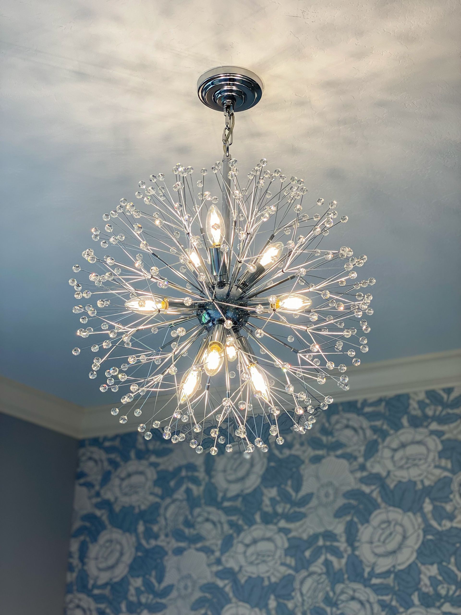 Shiny, starburst-shaped chandelier with crystal accents, hanging from a chrome ceiling fixture, in a room with floral wallpaper.