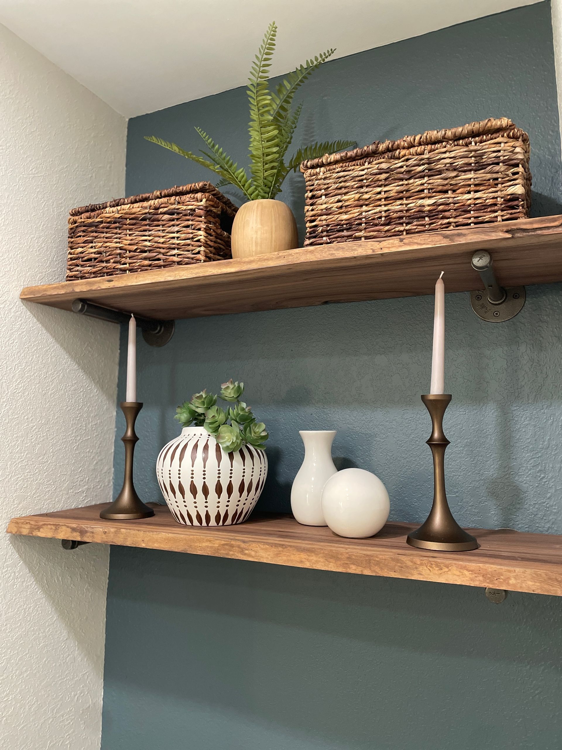 Two wooden shelves with decor against a teal wall: baskets, greenery, candles, and vases.