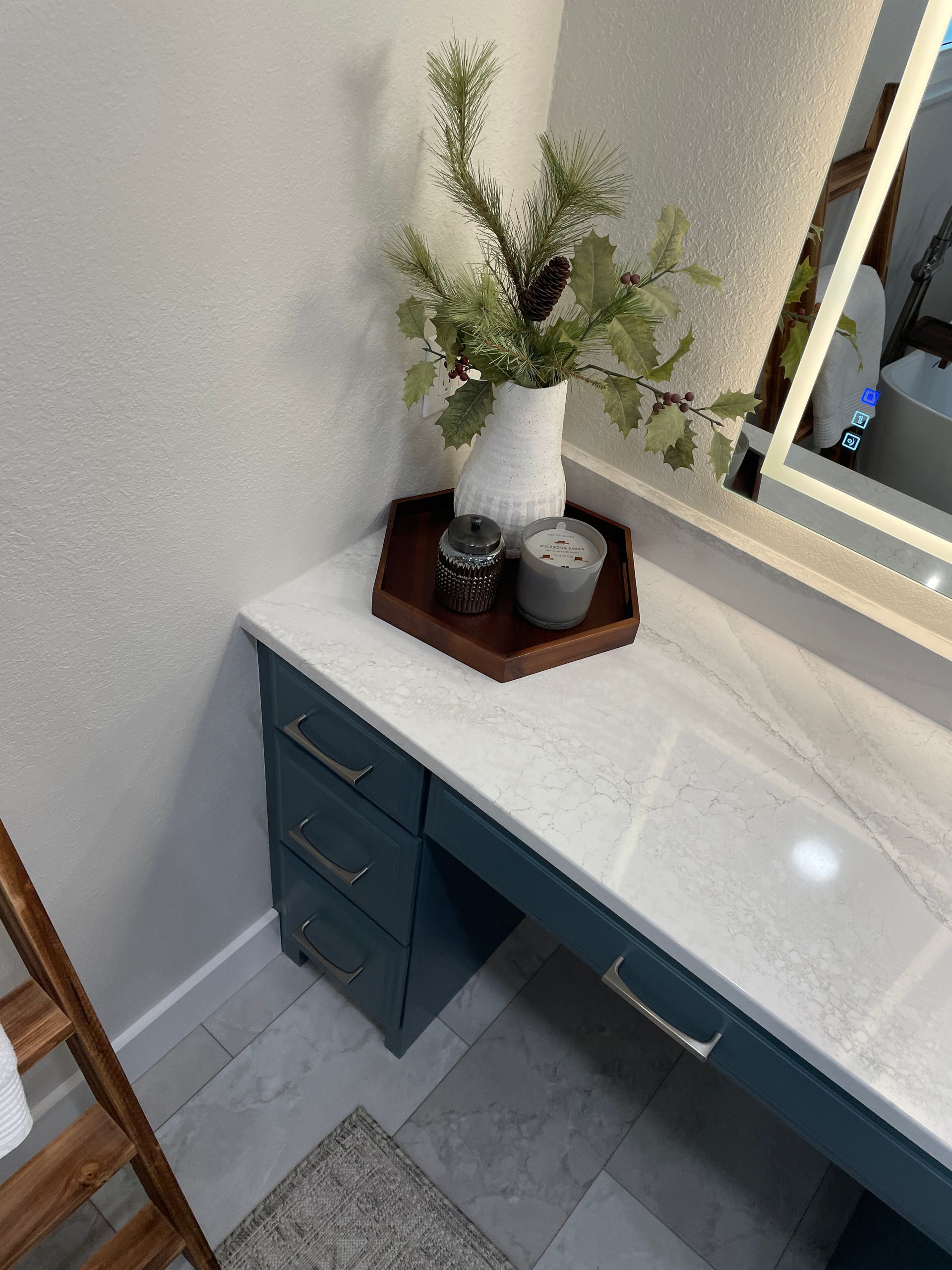 Vanity with blue cabinets, white countertop, vase with greenery, decorative tray, and lit mirror.