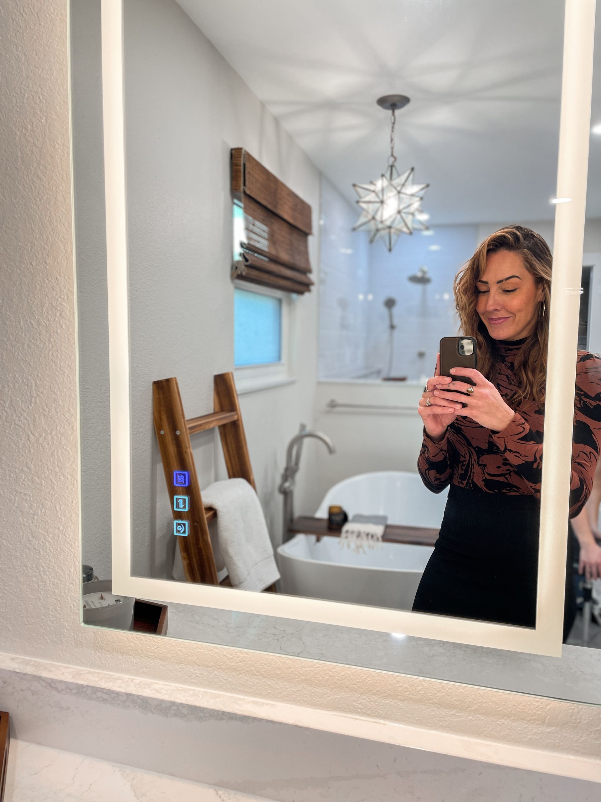 Woman taking a selfie in a modern bathroom mirror, smiling. The bathroom has a wooden ladder, bathtub, and star-shaped light.