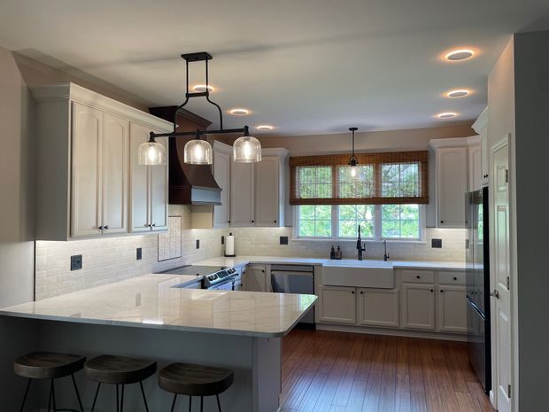 White kitchen with island, wooden stools, and dark light fixtures, with a wood floor and bamboo shade over window.