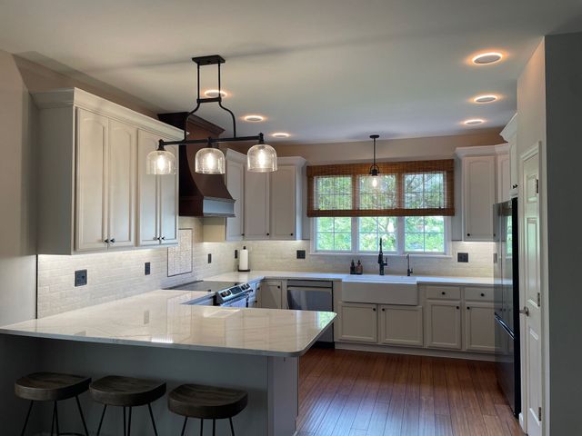 White kitchen with island, wooden stools, and dark light fixtures, with a wood floor and bamboo shade over window.