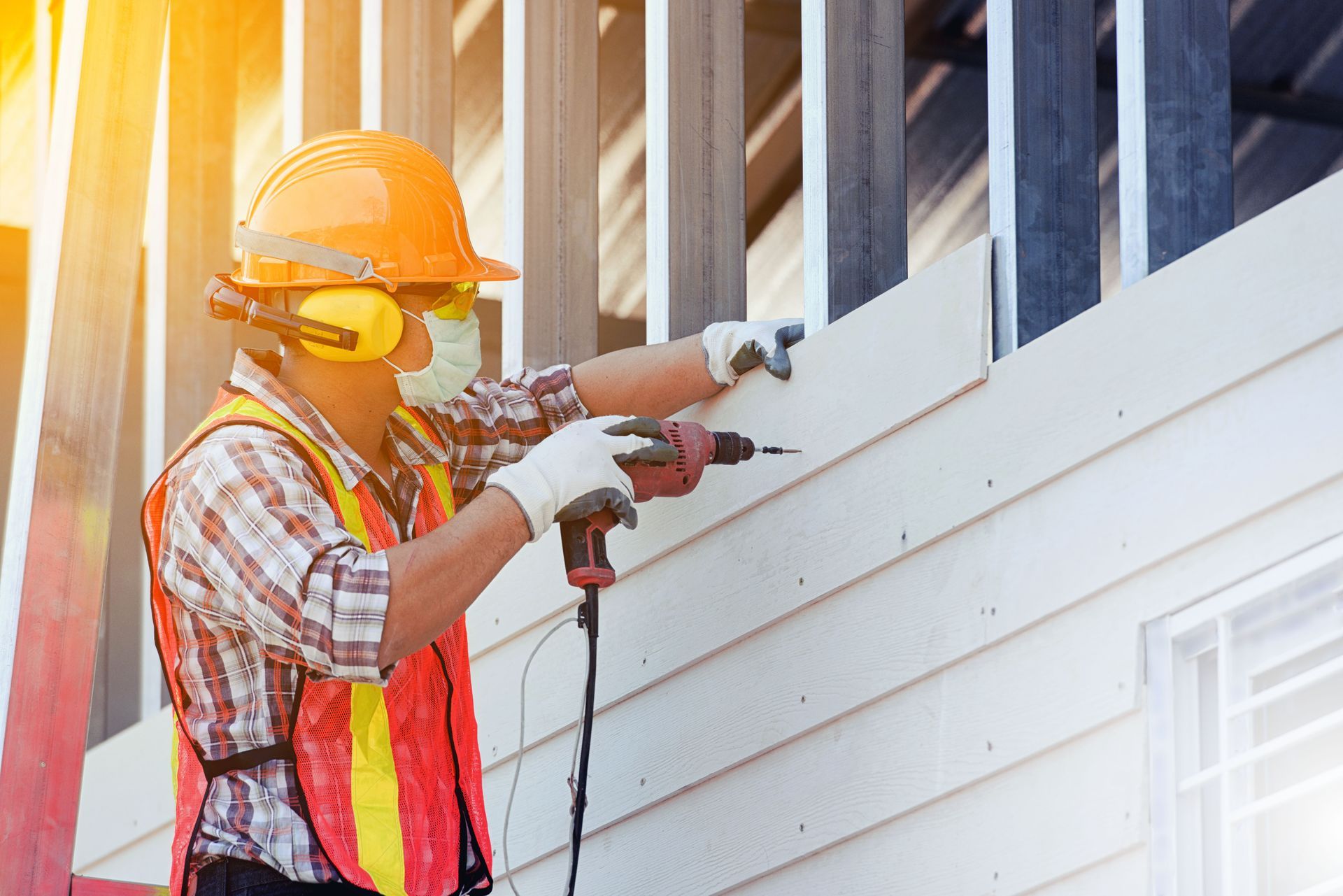 Worker in orange hard hat and safety vest spraying white paint on house siding