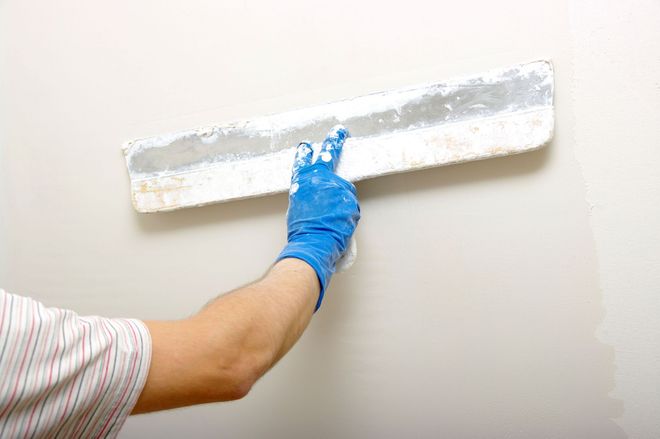 A person wearing a blue glove uses a metal trowel to apply white spackle or joint compound to a wall.