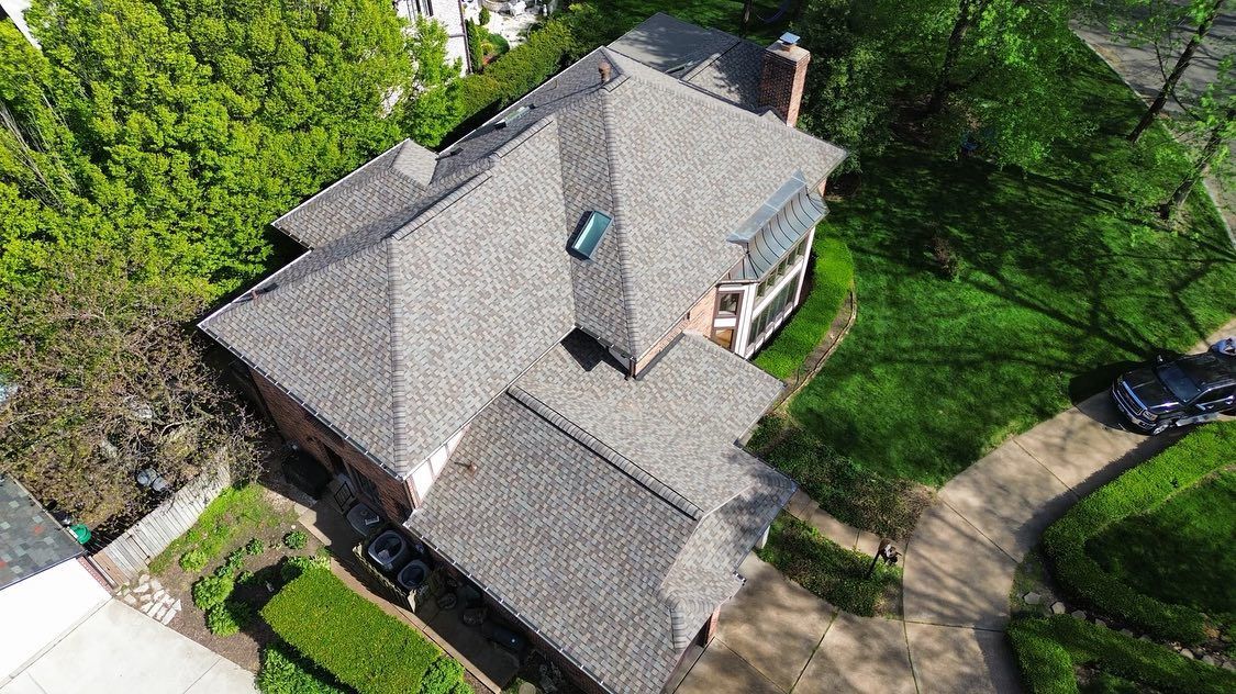 Overhead view of a house with a gray roof, surrounded by green grass and trees, and a driveway.