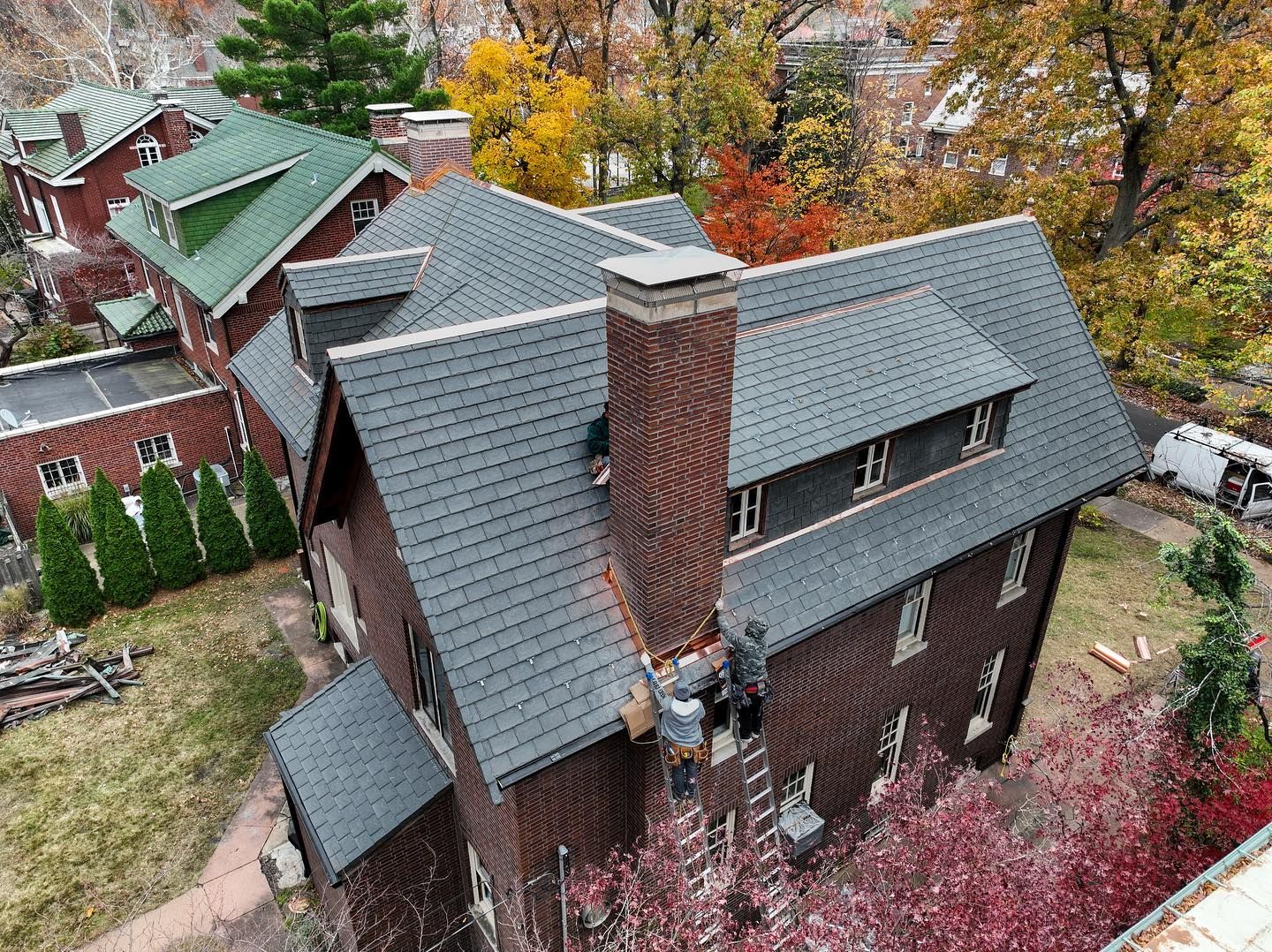 A multi-story brown house with a dark gray roof and chimney, autumn trees surround it.