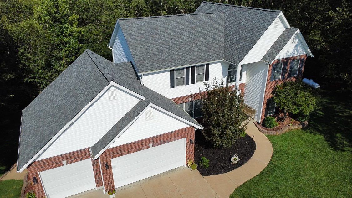 Two-story house with gray roof, white siding, and brick accents; surrounded by trees and green lawn.