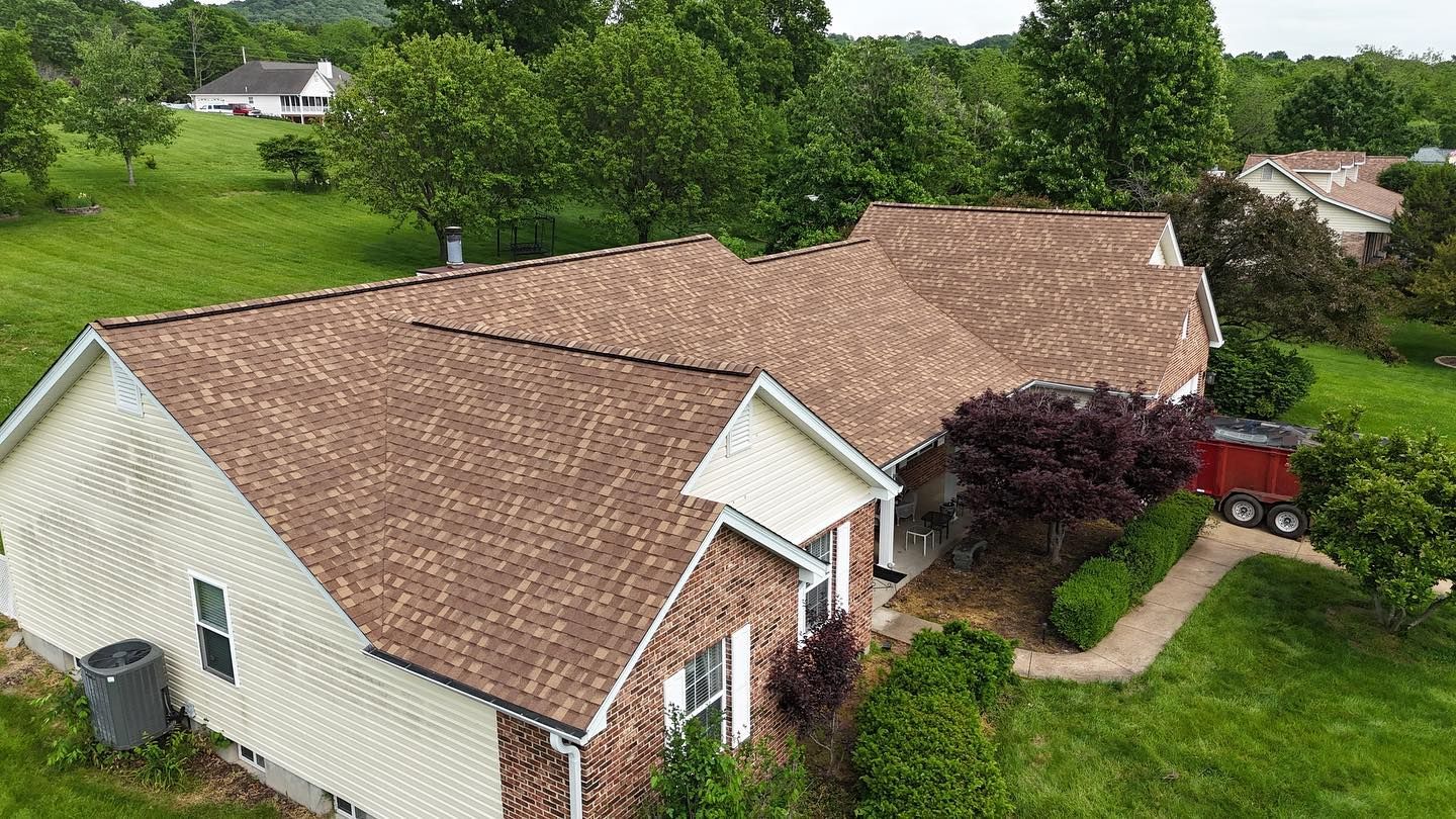 Brown roofed house on a green hill with trees, and a small red trailer.