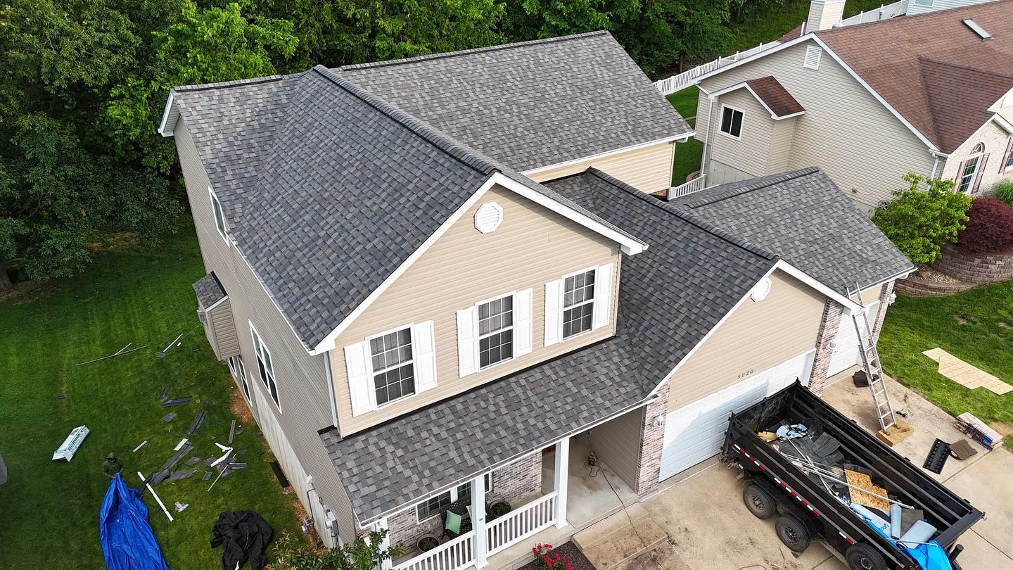 House with new gray roof, dump truck in driveway, green lawn.