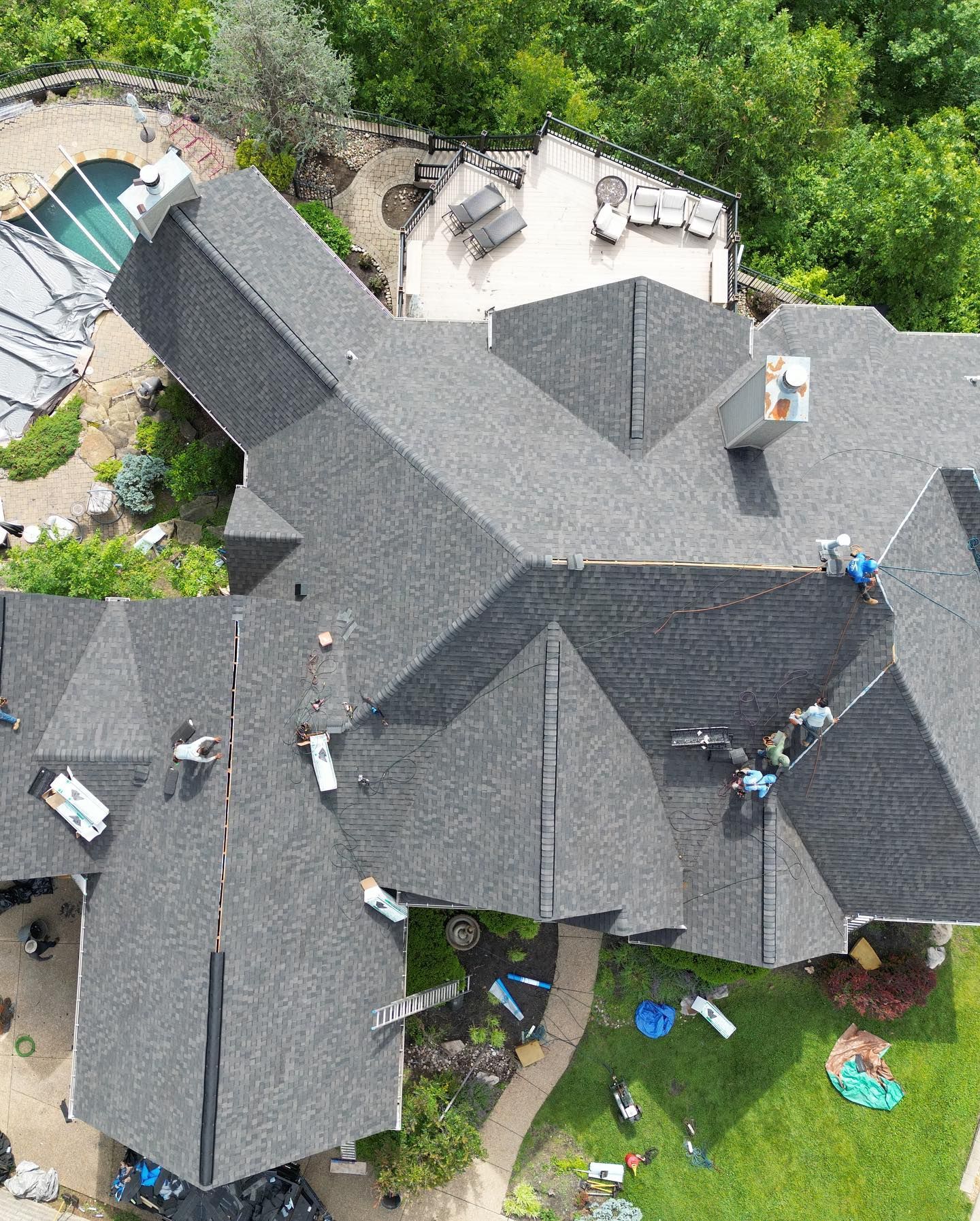 Workers on a dark shingled roof, trees surround the house with a pool visible.