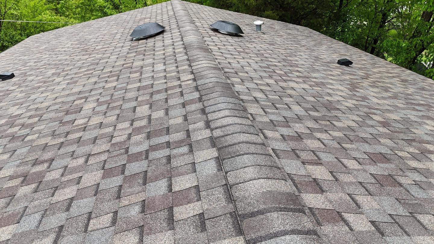 Close-up view of a brown and grey shingled roof with a vent and trees in the background.