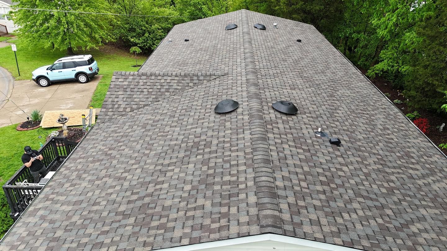 Overhead view of a house roof with brown shingles, black vents, and a blue car parked in the driveway.