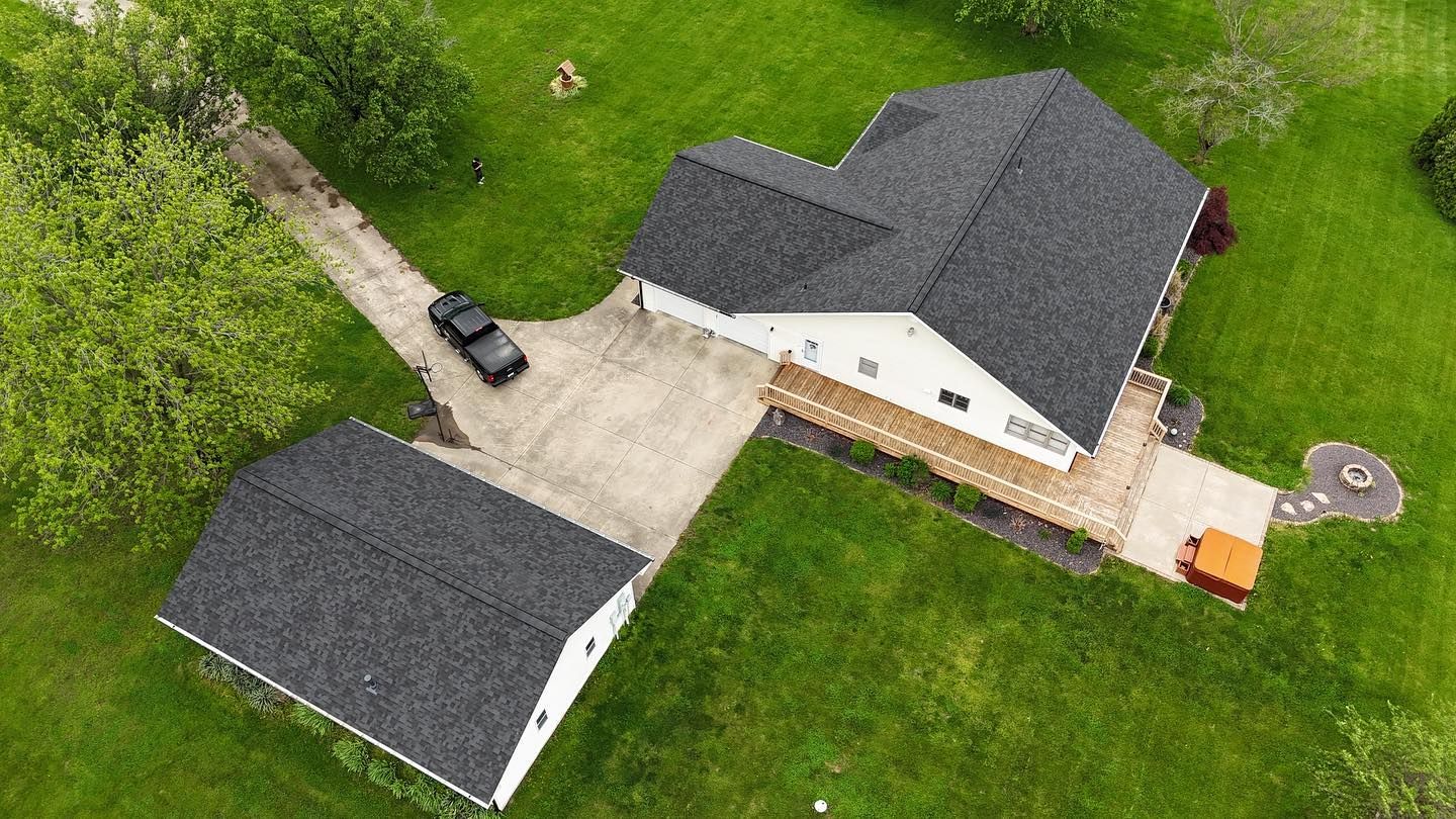 Aerial view of a white house and detached garage with gray shingle roofs, surrounded by green grass and a concrete driveway.