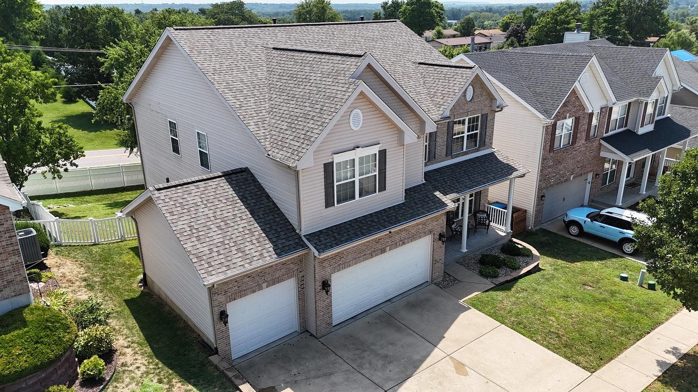 Two-story beige house with a dark roof and two-car garage, viewed from above, in a suburban neighborhood.