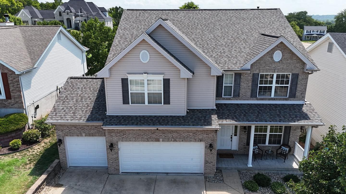 Two-story house with gray roof, tan siding, brick accents, and two-car garage on a sunny day.