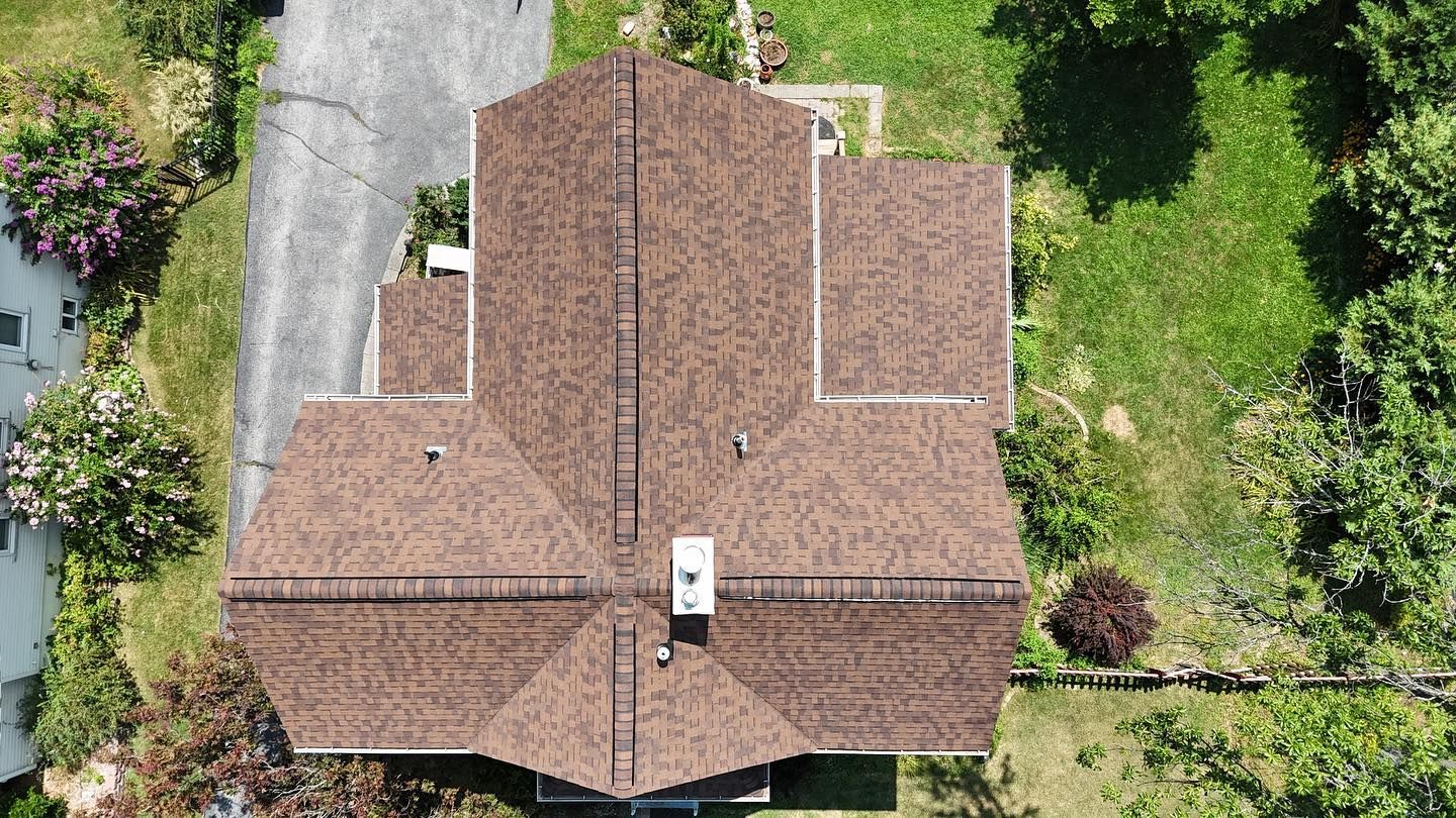 Overhead view of a house with a brown shingle roof, surrounded by green grass, trees, and a driveway.