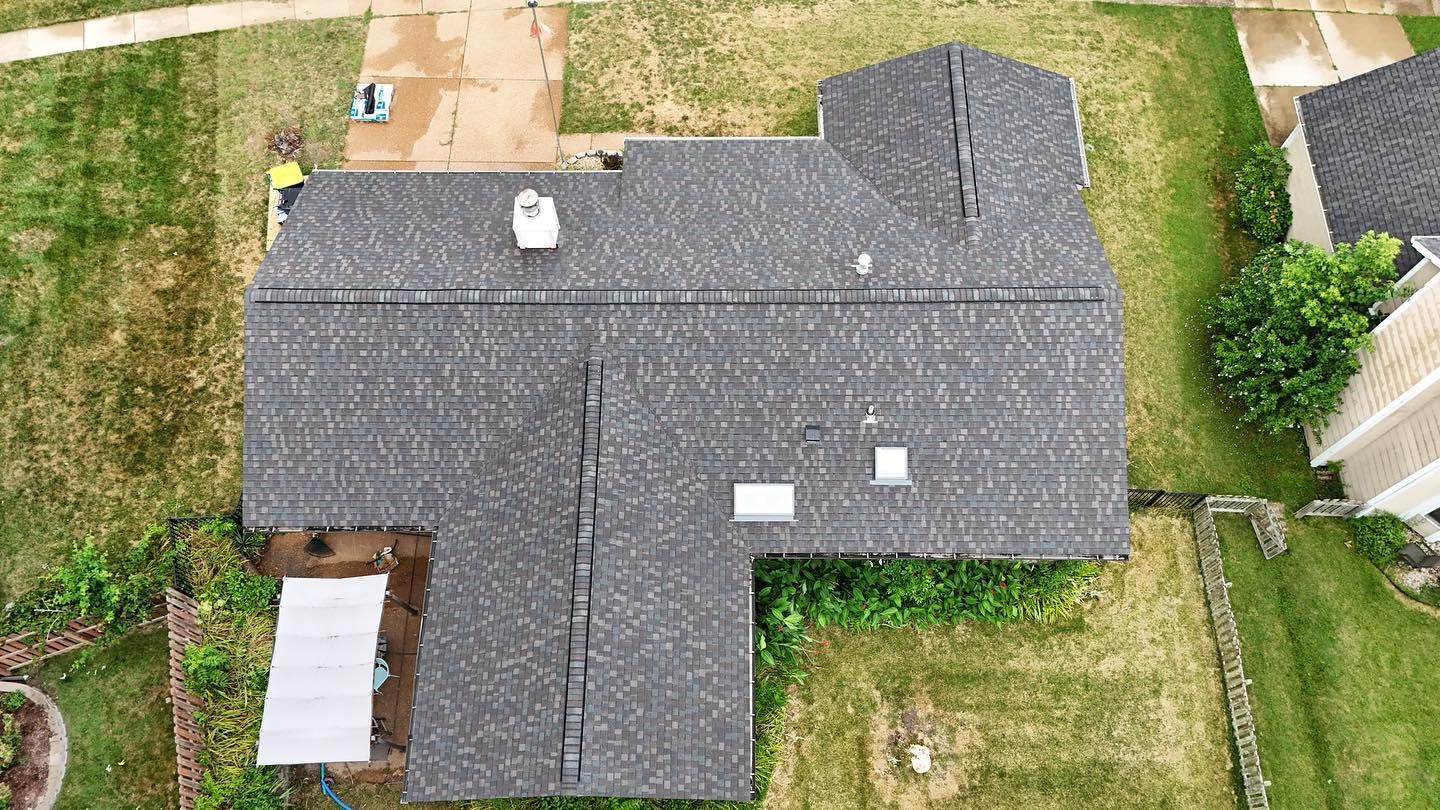 Overhead view of a dark gray shingled roof with a chimney and skylight. Surrounded by green grass and neighboring house.