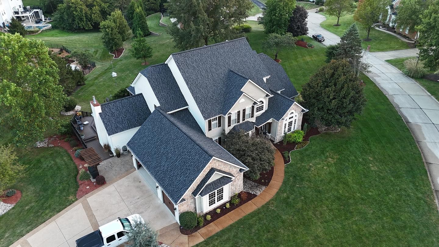 Aerial view of a two-story house with dark gray roof, beige siding, and green lawn. Driveway with a white truck.
