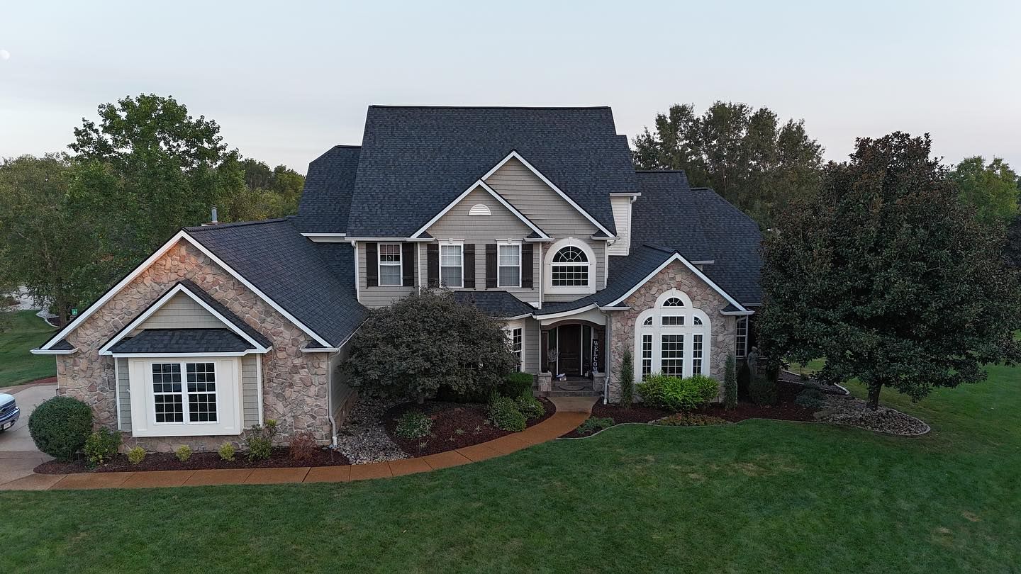 Large stone and grey roof house with lush green lawn and trees.