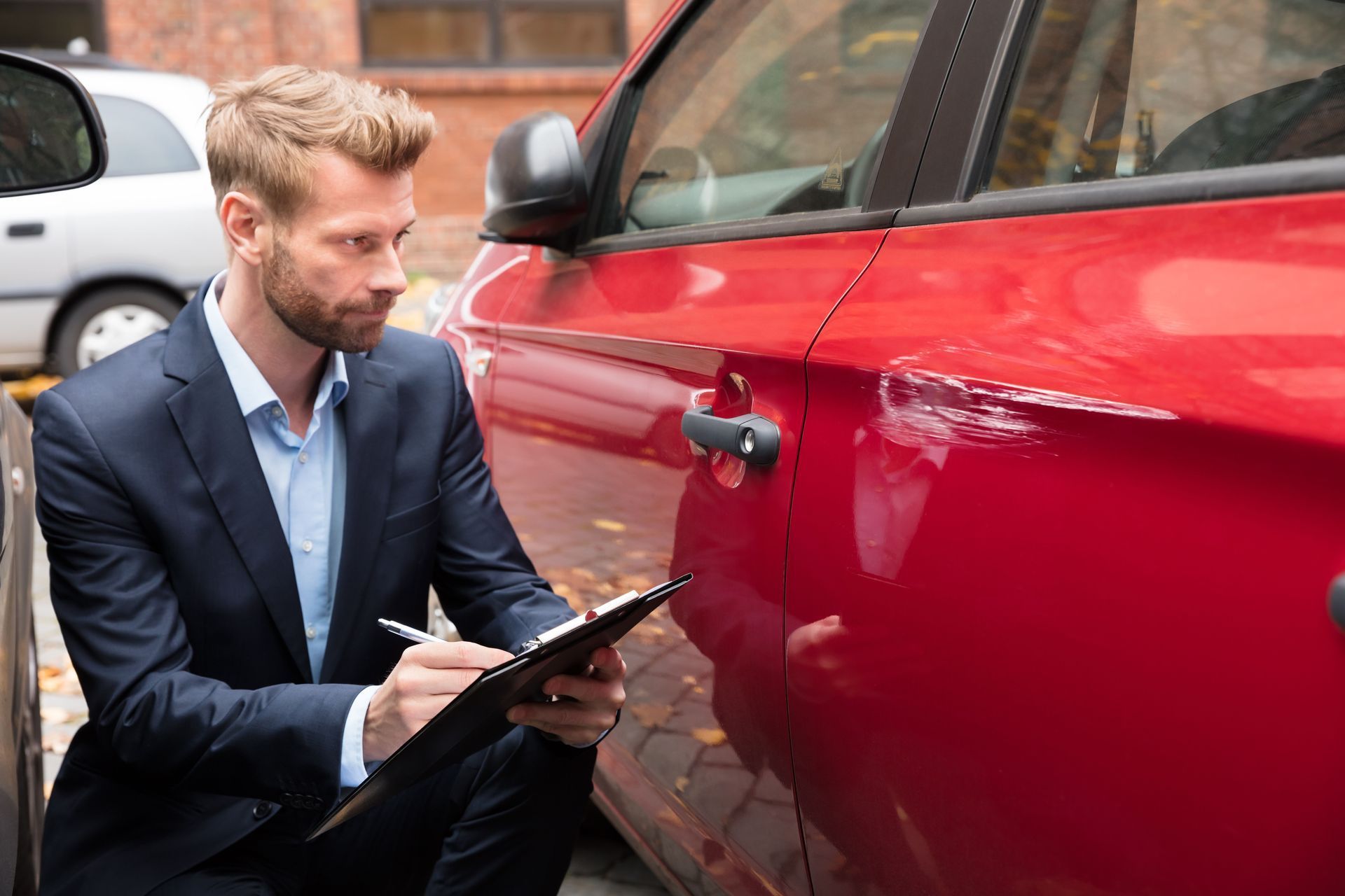 Man in suit examining a red car, taking notes on a clipboard outdoors.