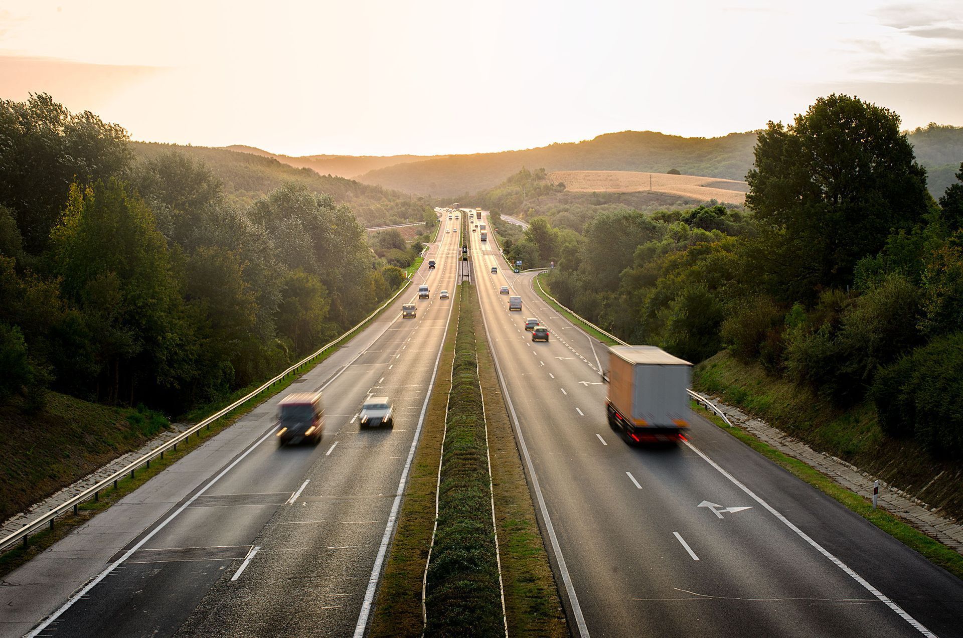 Highway with vehicles driving between green trees and hills at dusk.